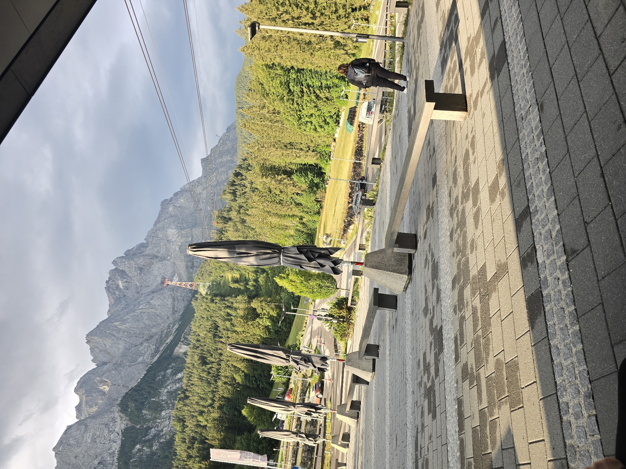 In the foreground, a paved plaza with a checkered pattern of light and dark gray stones leads to a view of a mountain range. Several large, dark umbrella structures, folded and covered, stand on concrete bases. A wooden bench with concrete supports is positioned in front of some of these umbrellas. A lone individual, wearing dark clothing and a backpack, walks away from the camera on the plaza. In the mid-ground, a road curves through a green, tree-covered hillside. A few cars are parked along the road. Further up the hillside, a ski lift tower with red accents is visible. The background is dominated by rugged, gray, rocky mountains under a cloudy sky. The scene appears to be the base area of a cable car or ski lift in Grainau, Germany, likely during the off-season or early morning, given the folded umbrellas and sparse activity.