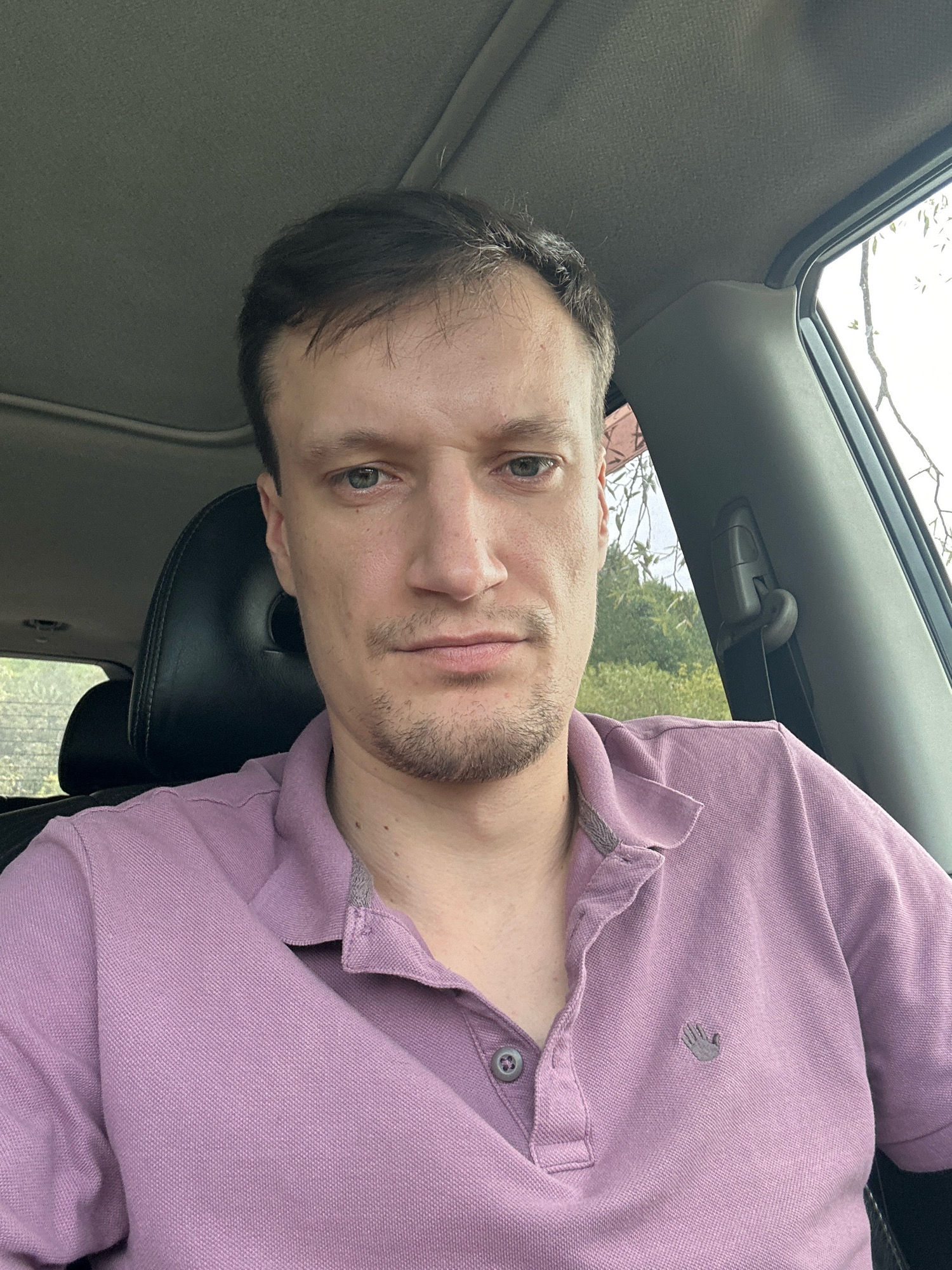 A man, appearing to be in his late 20s or early 30s, is pictured from the chest up, sitting in the driver's seat of a car. He has short, dark brown hair with some lighter strands, and light green eyes. His facial expression is neutral, and he has a light stubble. He is wearing a light purple polo shirt with a small grey logo of a hand on the left breast. The car's interior is visible, with the headrest of the passenger seat behind him and a glimpse of the car's ceiling. Outside the car window, trees and foliage are visible, suggesting an outdoor setting, possibly in a rural or suburban area. There is no specific indication of the location being Makiv, Ukraine.
