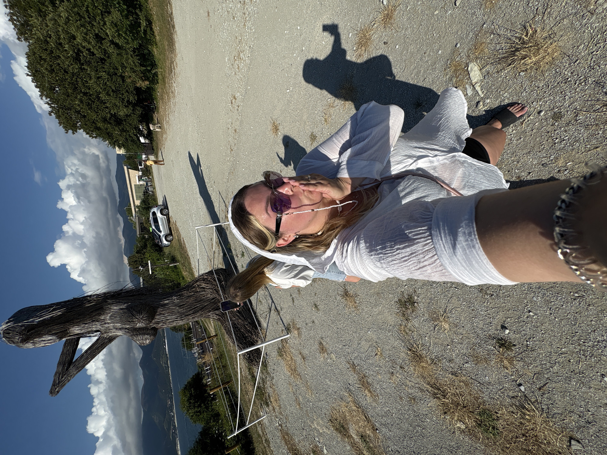 The image captures a sunny outdoor scene near the Black Sea coast in Kabardinka, Russia. In the foreground, a woman is taking a selfie with her left arm extended, holding a phone. She is wearing a white, flowing garment, dark shorts, and black sandals. Her right hand is held to her mouth, and she is wearing sunglasses and a pearl necklace with a beaded chain attached to her glasses. To the left of the woman, a large, abstract wooden sculpture resembling a human figure stands prominently. The sculpture is made of intertwined branches and appears to be weathered. Behind the sculpture, another woman with long blonde hair is visible, holding a phone and seemingly taking a picture. She is wearing a light-colored top and denim shorts. In the background, a body of water, likely the Black Sea, stretches out to the horizon, with mountains visible in the distance. A few buildings and trees line the coast. A white fence separates the viewing area from the cliff edge. The ground is covered in gravel and dry grass. Other individuals and a white car are visible further in the background, suggesting a public or tourist area. The sky is blue with scattered white clouds.