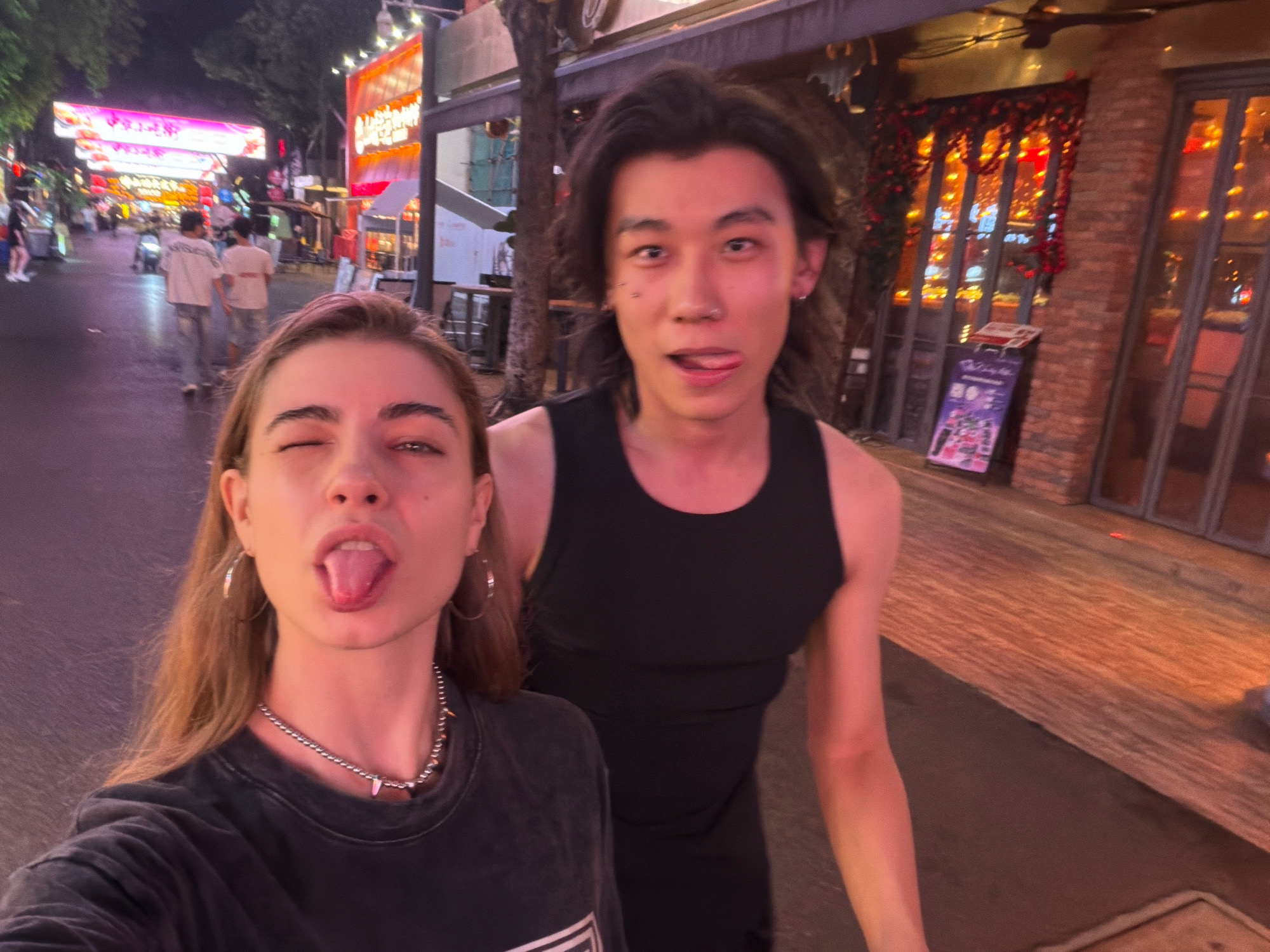 A young Asian man wearing a black tank top and a young Caucasian woman wearing a dark t-shirt are taking a selfie on a city street at night. The woman is in the foreground, sticking her tongue out and winking. The man is behind her, also sticking his tongue out. The street is lined with shops and restaurants with neon signs. The man has shoulder-length dark hair and several piercings on his face. The woman has long brown hair, hoop earrings, and a silver necklace. In the background, two people wearing white t-shirts and jeans walk away from the camera. The overall scene is brightly lit by the shop signs and the lights from inside the buildings, indicating a bustling urban environment. The location appears to be Chancheng District, China, based on the visual cues of the shops and signage.