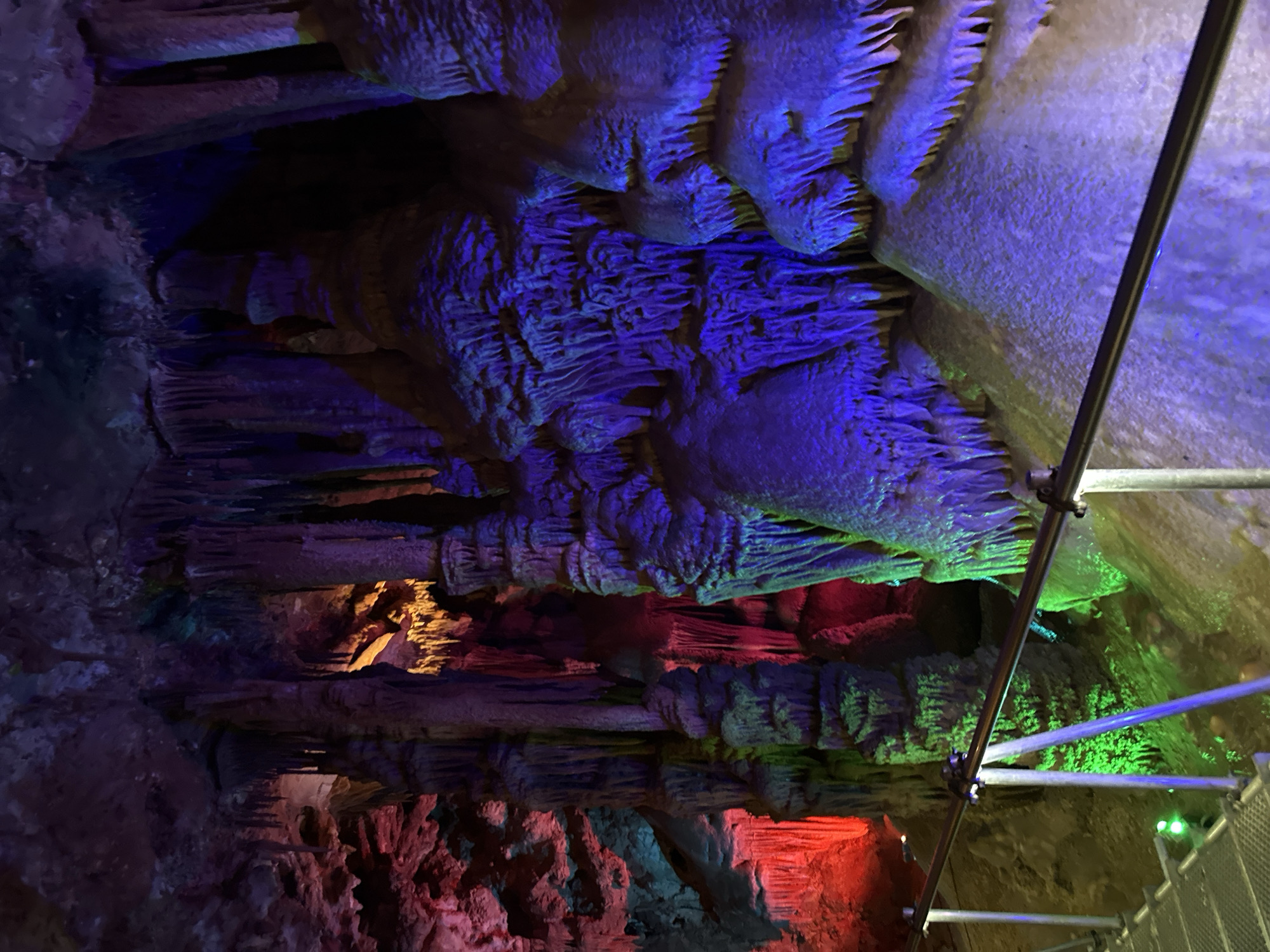The image shows the interior of a cave, likely located in Belo pole, Bulgaria. The cave walls are textured with formations like stalactites and stalagmites. The cave is illuminated with colored lights, primarily in shades of blue, green, red, and yellow, highlighting the textures and shapes of the cave formations. A metal scaffolding structure is visible on the right side of the image.