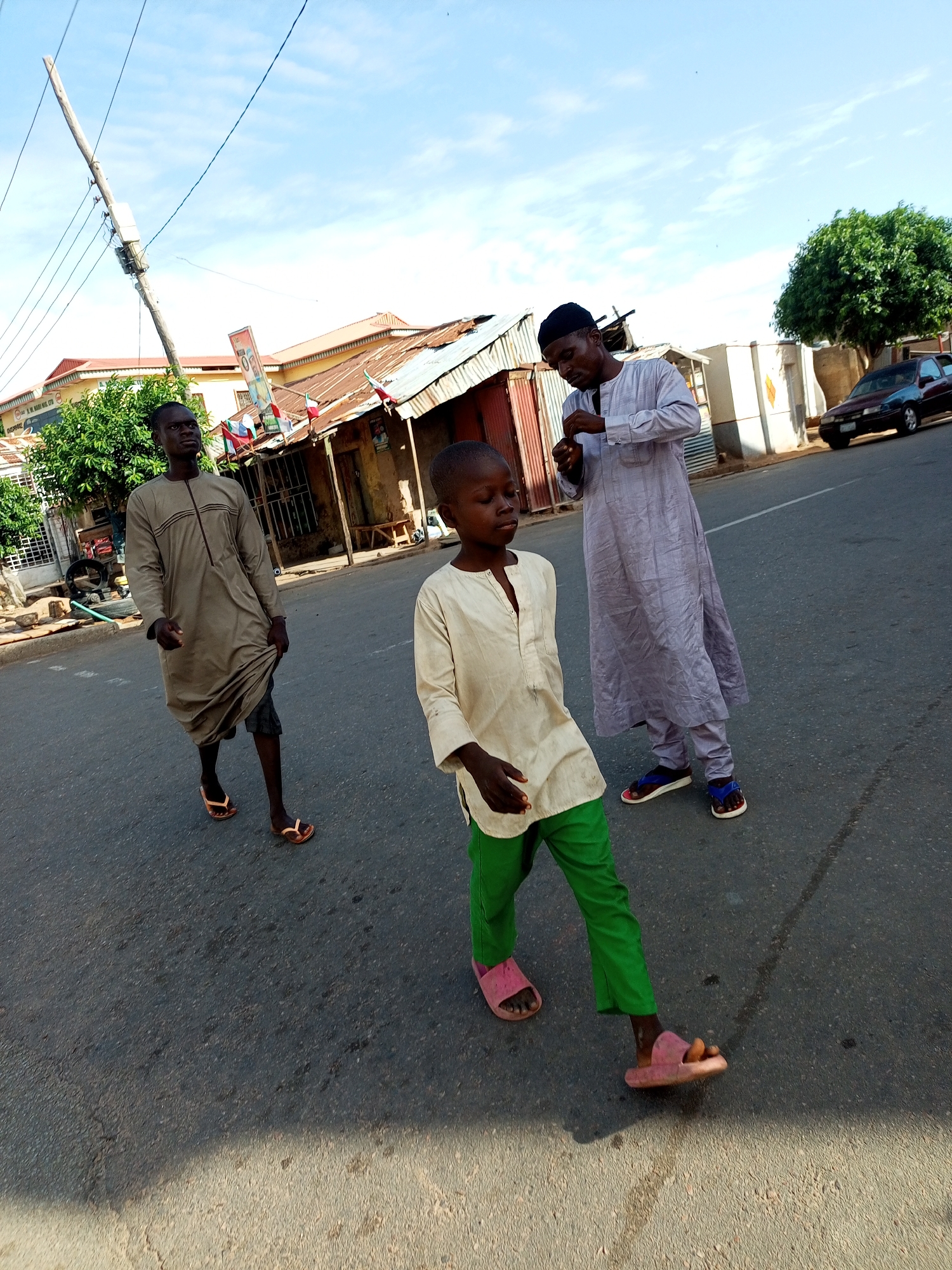 Three people, two men and a young boy, are walking down a paved road in what appears to be a town in Arawa, Nigeria. The setting includes buildings with corrugated metal roofs and some vegetation. The sky is blue with scattered clouds, suggesting it is daytime, likely late morning or early afternoon.
The man on the left is wearing a long, light brown robe over dark shorts and open-toed sandals. He is looking to his right. In the center, a young boy is walking ahead of the man on the left. He is wearing a light-colored shirt and bright green trousers, with pink sandals. He appears to be looking down and slightly to his left. The third person, a man on the right, is wearing a light purple robe and matching trousers, also with open-toed sandals. He is looking down and to his left, possibly at his phone or something in his hand.
In the background, there are several structures, some appearing to be shops or houses with weathered facades. There are also flags visible on one of the buildings, and a car is parked on the right side of the road. A utility pole with electrical wires is prominent on the left side of the image. The overall atmosphere seems casual, with the individuals walking along the road. There is no specific event depicted, but rather a snapshot of people moving through a public space.
