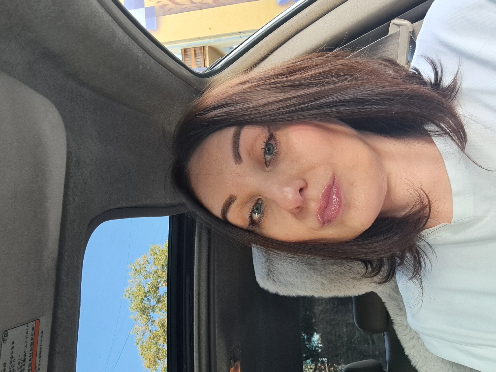 A woman is taking a selfie in a car. She has dark brown hair and is wearing a white t-shirt. She is wearing lip gloss. Through the sunroof, a tree and blue sky are visible. The car's interior, including the sunroof, seats, and part of the dashboard, are also visible. A building with a yellow facade and balconies can be seen through the car window. The image is taken from a slightly low angle. There is no discernible location in the city of Irkutsk, Russia.