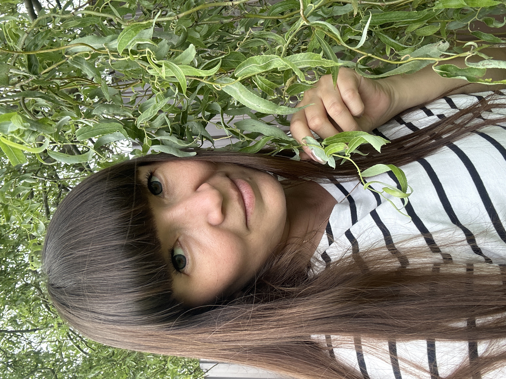 A woman with long, dark brown hair and bangs stands outdoors in front of a weeping willow tree, its branches partially obscuring her face. She is wearing a black and white striped t-shirt and a gold chain necklace. Her left hand is visible, reaching out to touch the leaves of the tree. The background consists of the tree's foliage and a blurred fence. The setting appears to be a residential area in the village of Sredneye Kechevo, Russia.