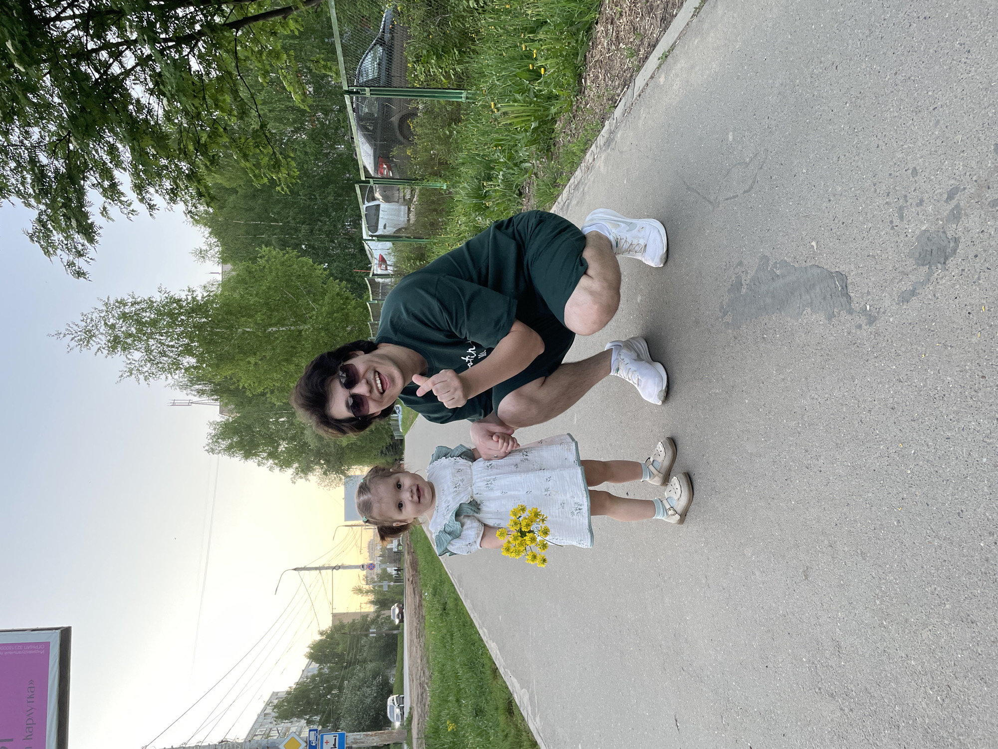 The image shows an adult male and a young girl on a sidewalk in Izhevsk, Russia. The man is squatting, wearing a dark green t-shirt, dark shorts, sunglasses, and white sneakers. He is giving a thumbs-up sign. The girl is standing next to him, holding yellow flowers, wearing a light-colored dress, white socks, and light-colored shoes. A road with buildings and trees is visible in the background. There is also a chain-link fence visible, and what appears to be vehicles behind it. Street signs, including one in Russian, are visible. A large advertising banner is on the left side of the image. The sun is setting, indicating late afternoon or early evening.