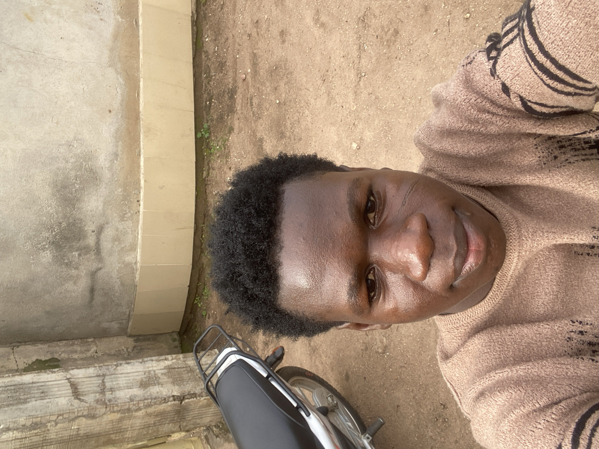 A close-up selfie captures a young male individual, appearing of African descent, looking directly into the camera in Mararaba, Nigeria. He has short, dark, curly hair and visible facial scars on his left cheek. He wears a light brown knitted sweater featuring dark, possibly black, striped patterns on the sleeves. His forehead and nose reflect light, suggesting a sheen on his skin. To the left and slightly behind him, the rear portion of a dark-colored motorcycle with a silver rear rack is partially visible. The ground is a light brown, unpaved surface with scattered small green plants. The background features a weathered, light beige-grey concrete wall with a horizontal band of lighter-colored tiles along its base, and a textured concrete pillar with some green discoloration on the far left.