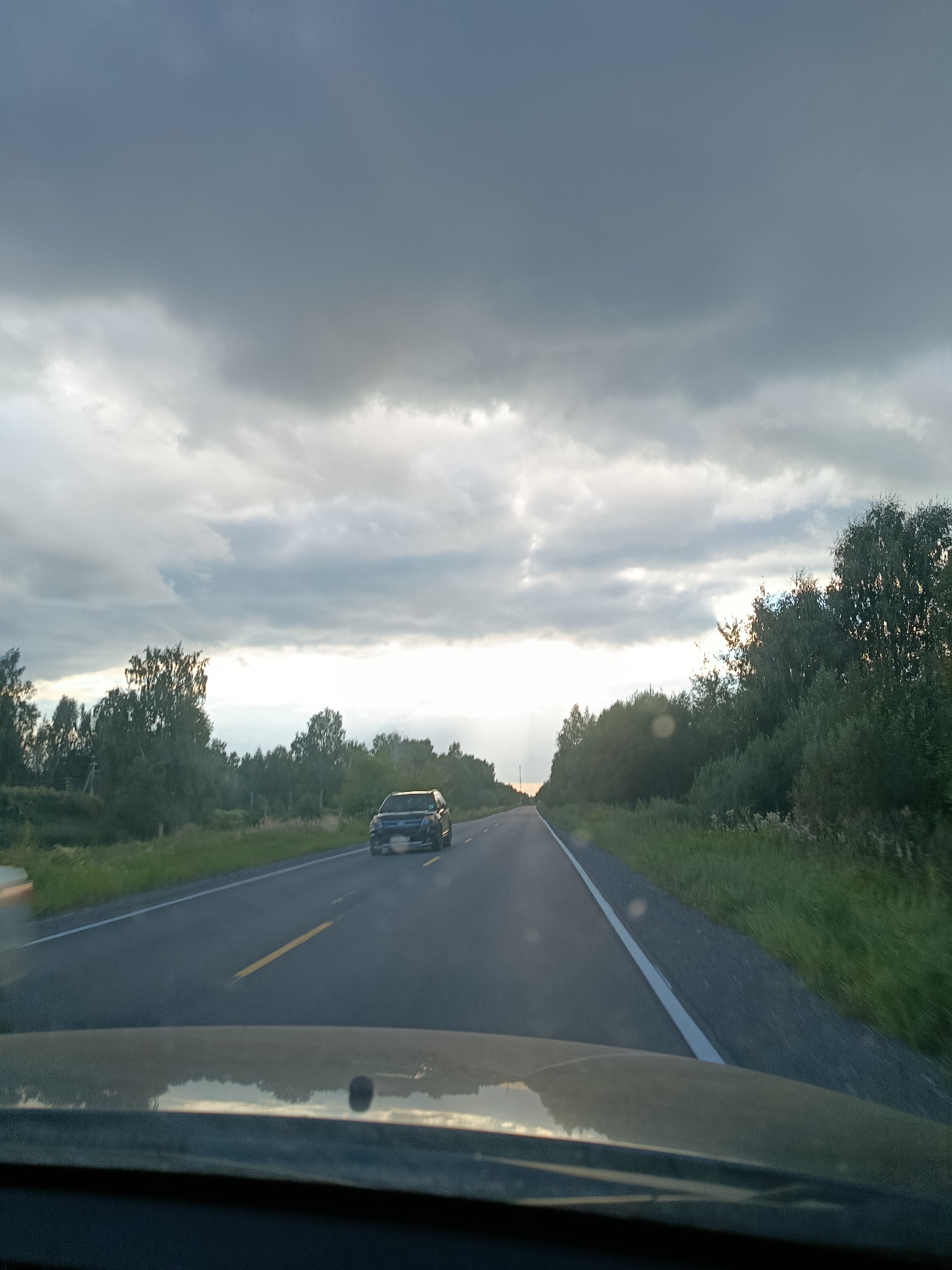 The image shows a dark, overcast sky with scattered clouds. A paved road stretches into the distance, bordered by green grass and trees on either side. A dark-colored car is driving on the road. The scene appears to be a rural or suburban area. There is no indication of the city of 1-Go Maya, Russia.