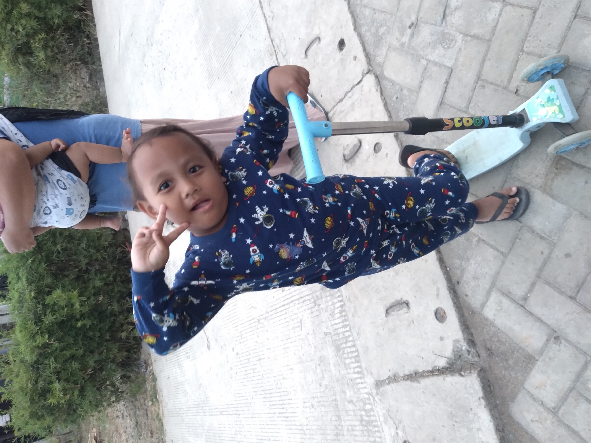 A young child in a blue pajama set with a space-themed print stands on a light blue three-wheeled scooter. The child is facing the camera, with their right arm raised in a peace sign gesture and their left hand on the scooter's handlebars. The child has dark hair and is looking directly at the camera with a playful expression. Behind the child, a person is holding another baby. The scene is outdoors, on a paved area with some vegetation in the background. The pavement is made of concrete slabs and interlocking bricks. The location is identified as Pandak, Indonesia.