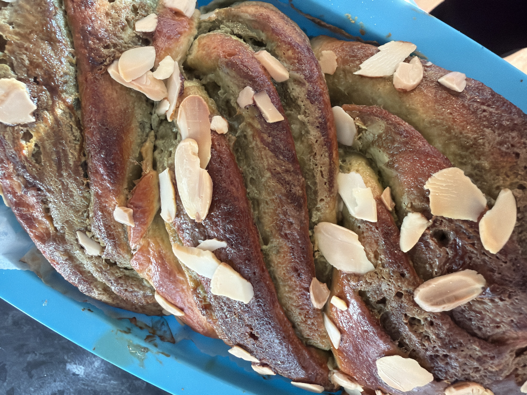 The image depicts a baked loaf of twisted bread in a blue baking pan. The bread is golden brown on the exterior, with a green-tinged interior. It is adorned with sliced almonds. The bread appears to be sitting on a counter. The location is not discernible from the image.