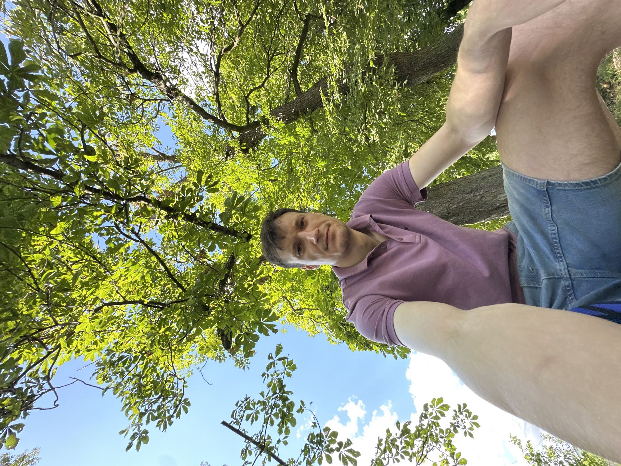 A low-angle, wide shot captures a man seated outdoors under a canopy of green trees. The man, appearing to be in his late 20s or early 30s with short brown hair and a light complexion, wears a pink polo shirt and denim shorts. He is looking directly at the camera with a neutral expression. His right arm rests on his thigh, while his left arm extends towards the camera, out of focus due to the close proximity. The background is dominated by the lush green foliage of trees, with patches of blue sky and white clouds visible through the leaves. The man is positioned on what appears to be grassy ground, with tree trunks visible behind him. The image is undated and does not contain any overt indications of the specific location in Makiv, Ukraine.