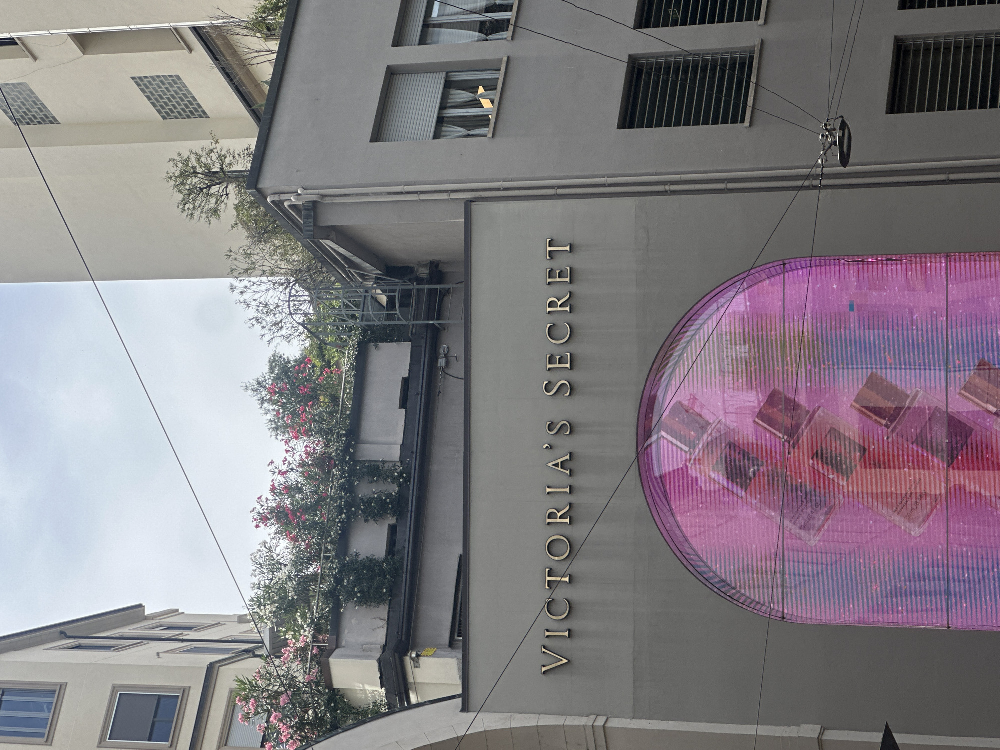 The image shows the facade of a Victoria's Secret store in Milan, Italy. The store has a large, arched digital display showcasing perfume bottles in shades of pink and purple. Above the store, there are balconies with blooming pink and white flowers, and a few small trees. The buildings surrounding the store are modern, with multiple windows and light-colored walls. The sky is visible in the background, with some clouds. There are no people visible in the image. The overall impression is one of a commercial street in a European city, with a focus on the storefront and its advertising.