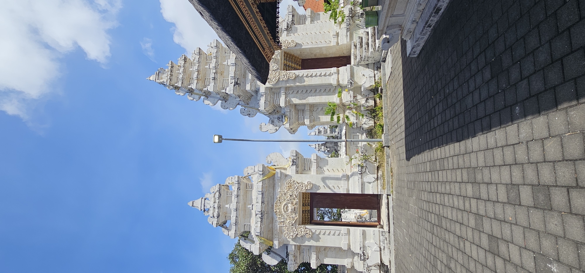 The image shows a traditional Balinese gate made of white stone, standing in Kuta Selatan, Indonesia. The gate features intricate carvings and a tiered roof, with a wooden door at its base. A lamppost stands nearby. The ground is paved with grey bricks. The sky is blue with some clouds.