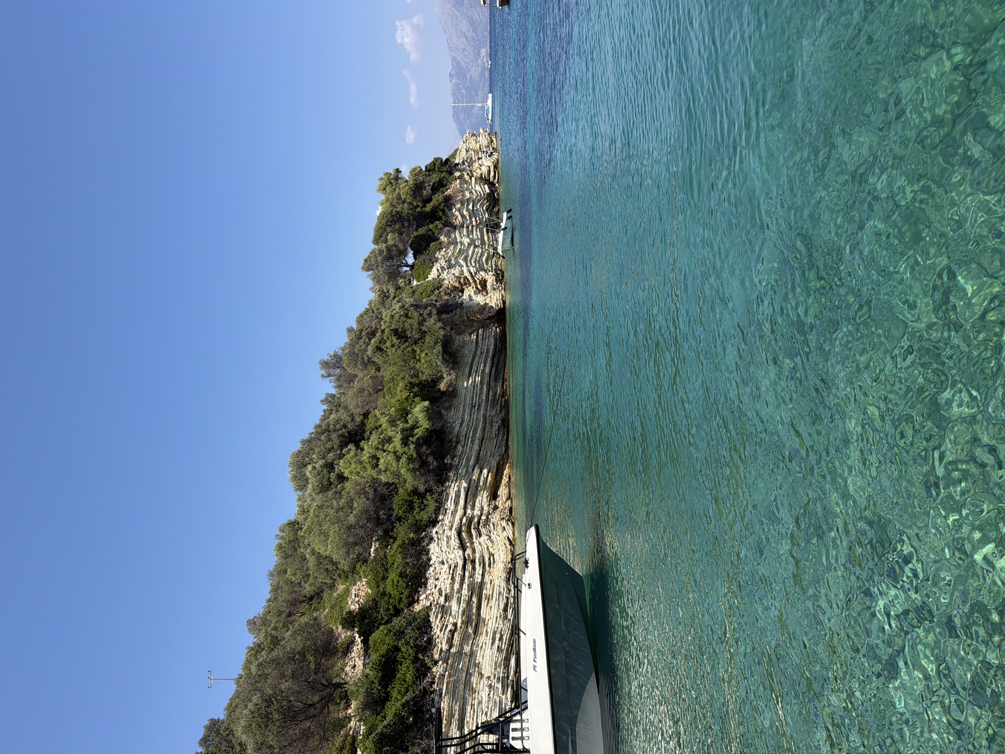 A powerboat is moored in the foreground in the turquoise waters off the coast of Kastos, Greece. The boat, a white and gray rigid inflatable boat, has a black metal frame structure. In the background, a rugged cliff face is covered with lush green vegetation, including olive trees. The rock layers of the cliff are clearly visible and appear to be light-colored and striated. The clear water allows for visibility of the seabed, which is rocky. Further in the distance, other boats, including a white sailboat, are visible on the water. The sky is clear and blue, indicating a sunny day. The scene depicts a tranquil coastal environment.