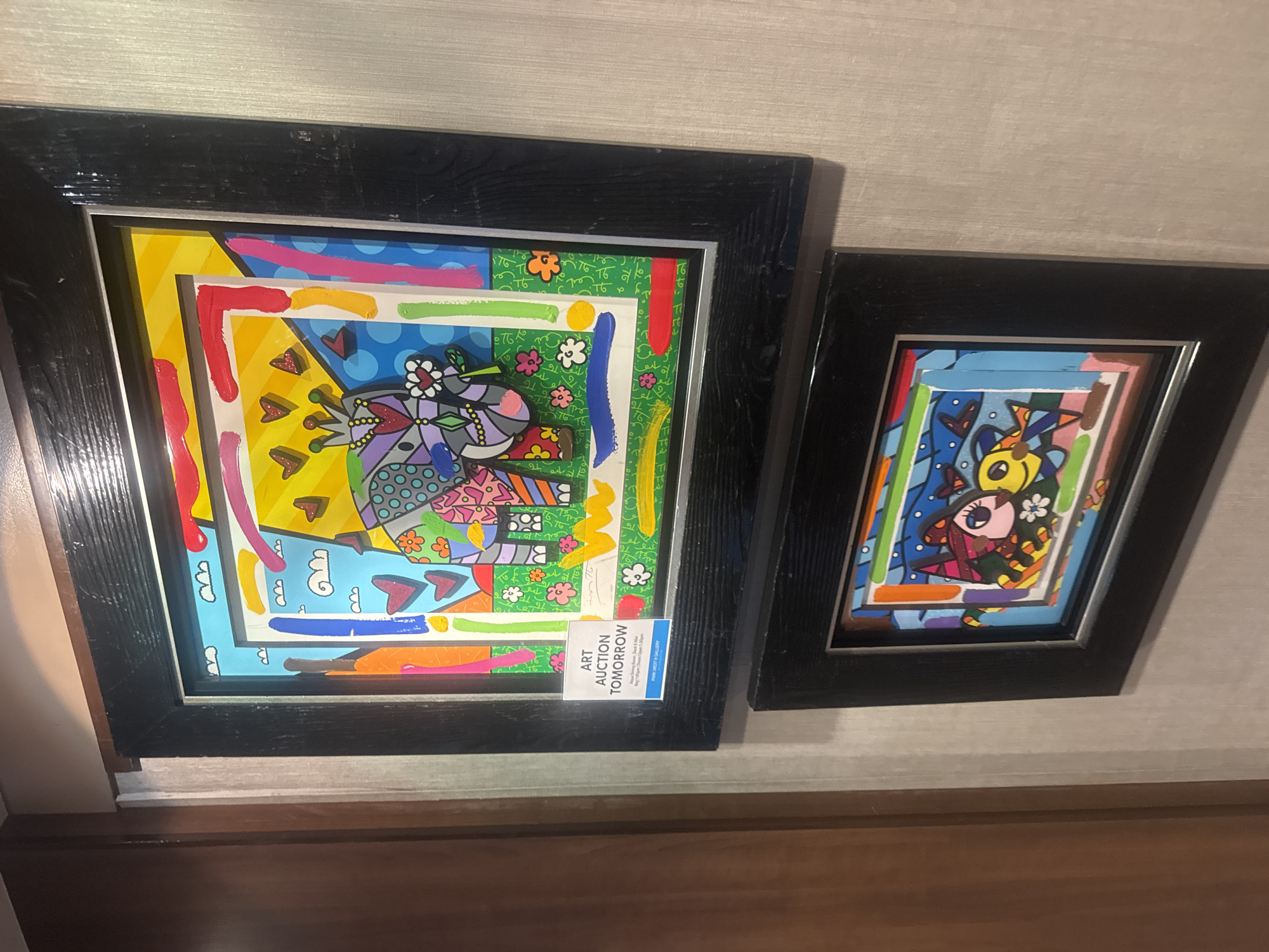 The image displays two framed pieces of art hanging on a wall with a textured, light-colored finish. The frames are black and appear to be made of wood. The left frame contains a white informational card that reads "ART AUCTION TOMORROW" with additional details about the auction location and time.
The artwork itself is brightly colored and features a whimsical, cartoonish style. The upper artwork depicts an elephant wearing a crown and holding a flower. The lower artwork features a stylized depiction of a girl with a witch's hat and a yellow fish. The art appears to be in the style of Romero Britto, known for his vibrant use of color and bold patterns.
The location is identified as Motril, Spain, and the context suggests this is a display within a gallery or event space related to an art auction.