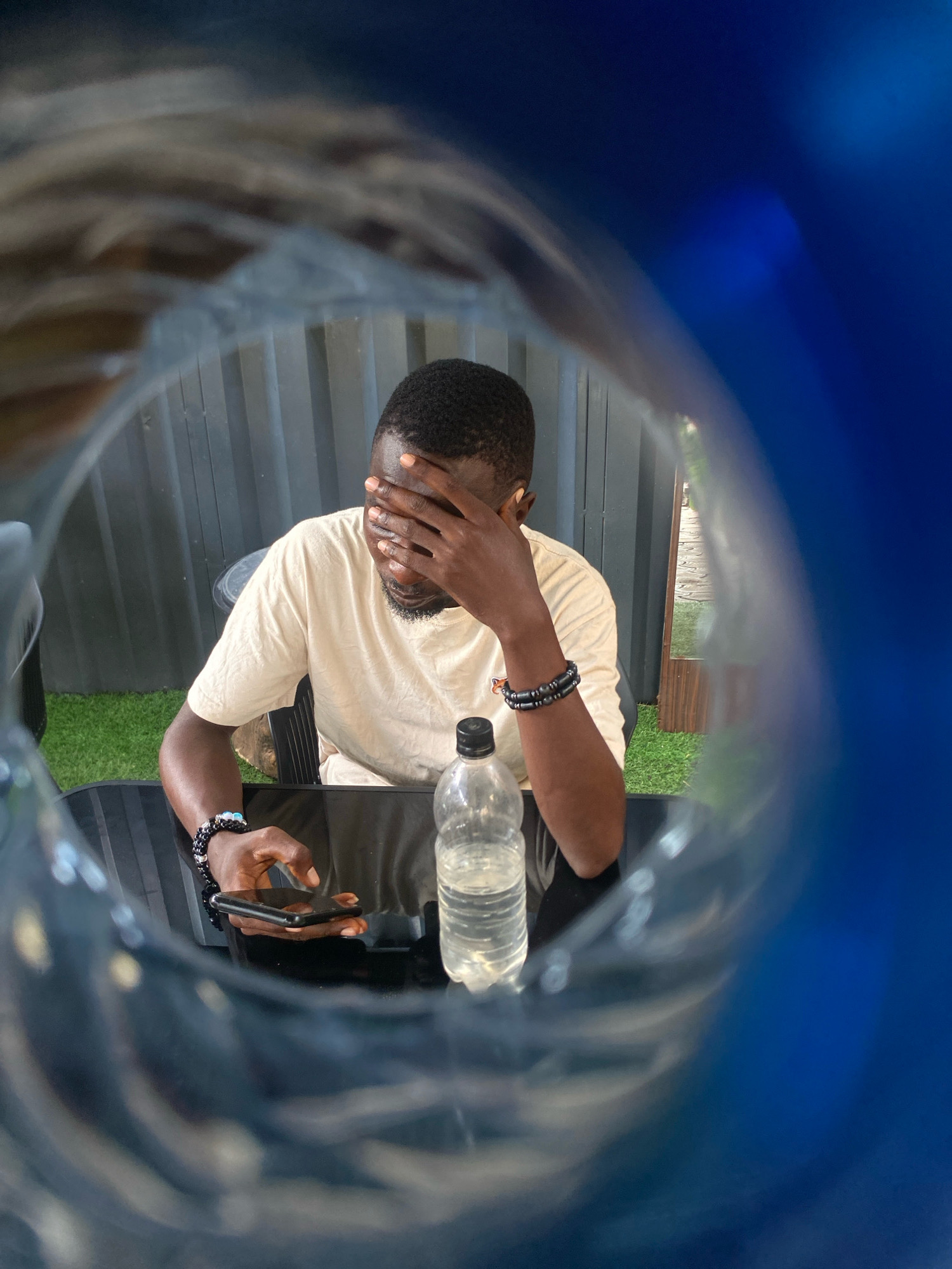 The image shows a man sitting at a black reflective table, holding his hand over his eyes, and looking down at a smartphone. He is wearing a light-colored t-shirt and two beaded bracelets on his left wrist. A bottle of water is on the table in front of him. The background appears to be an outdoor setting with a grey corrugated fence and some artificial grass. The perspective suggests the photo was taken through a translucent object, possibly a plastic bottle, with a blue tint. The scene is not identifiable as being in Barden Yana, Nigeria, or any other specific city.
