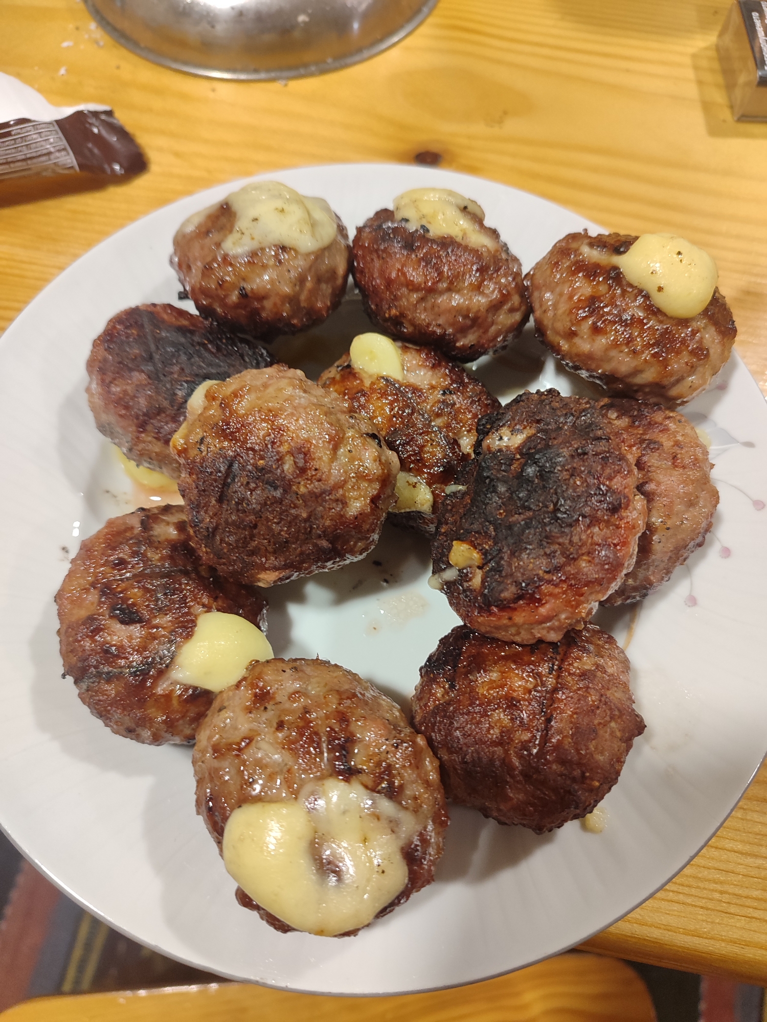 A top-down, close-up shot shows a white plate containing ten cooked meatballs, each topped with melted cheese. Some meatballs exhibit a darker, possibly burnt, exterior. A metallic lid is visible in the upper left corner. The surface beneath the plate appears to be a wooden table. A partial chocolate bar is also visible. The location is assumed to be Varshilo, Bulgaria. No people or actions are directly visible in the image.