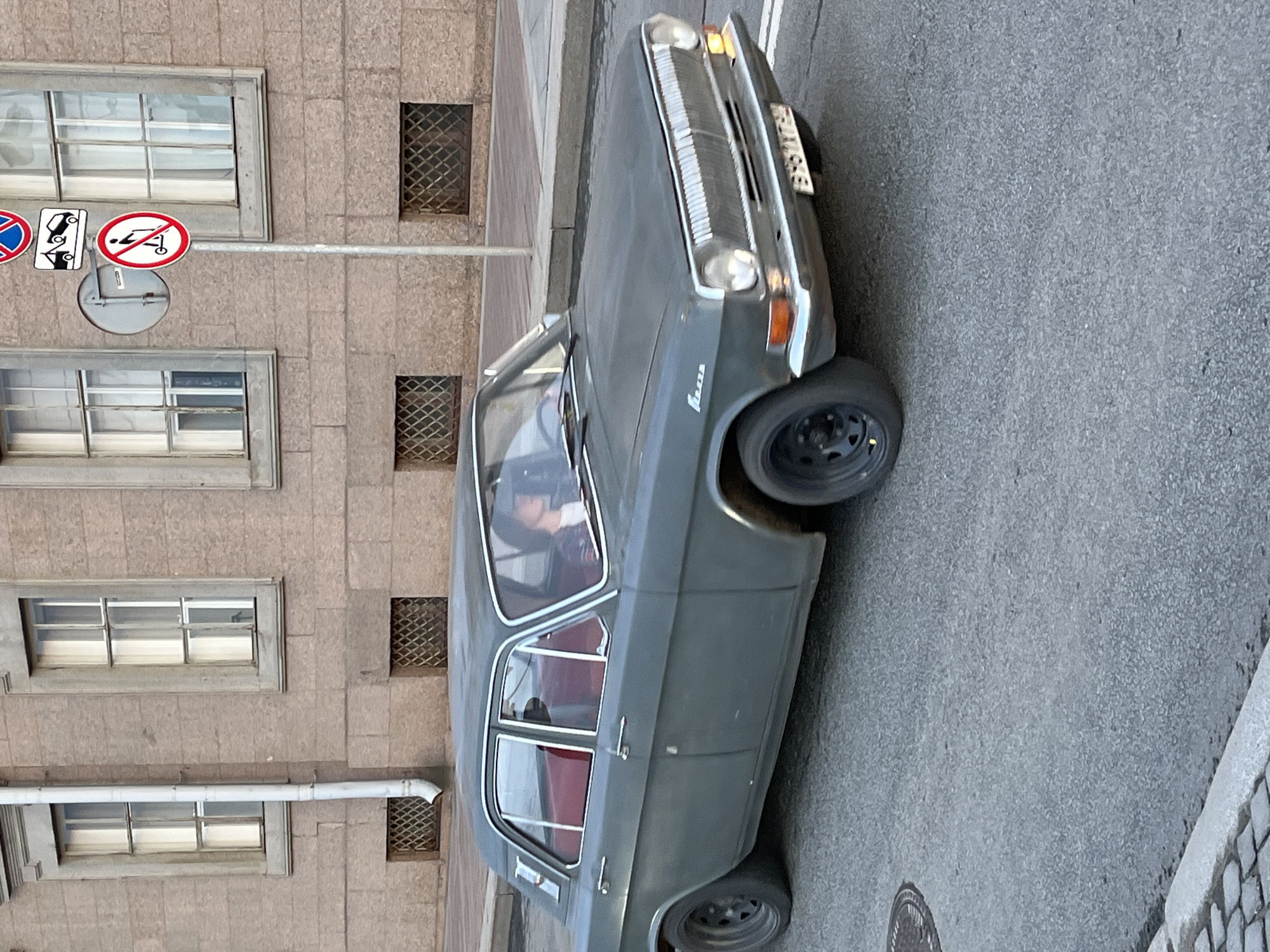 A grey GAZ-24 Volga car is parked on the right side of a grey asphalt road in Saint Petersburg, Russia. The car is oriented diagonally in the frame with its front end near the top and the back near the bottom. The side of a building is on the left with windows, stone detailing, and traffic signs. A person is visible inside the car behind the steering wheel. A manhole cover is visible on the street near the bottom.