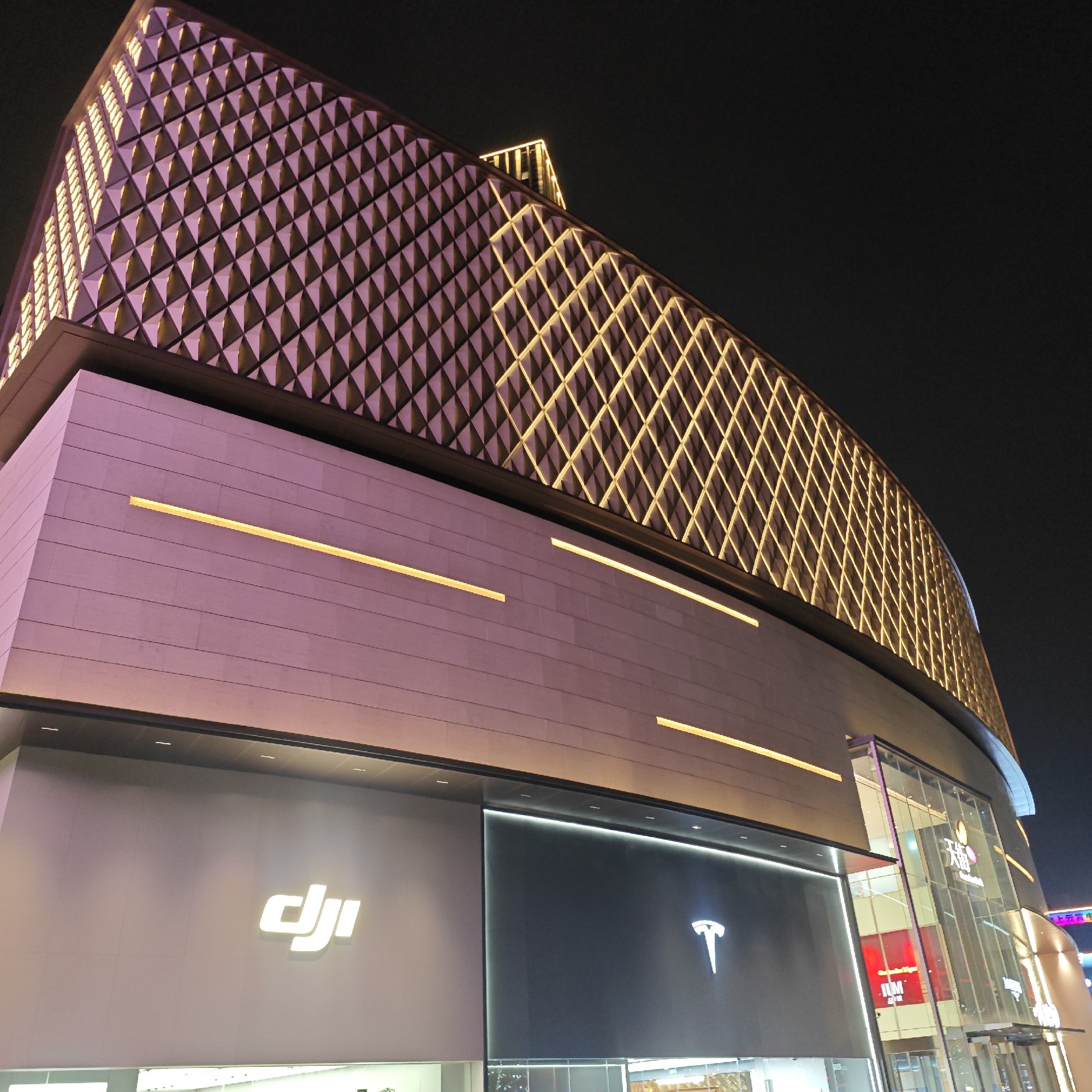 The image displays a modern commercial building at night, set against a dark sky in what appears to be a bustling urban environment, likely in Yuzhong District, China. The building's architecture is striking, featuring multiple layers and distinct lighting designs.
The uppermost section of the building exhibits a curved, textured facade covered in a geometric, diamond-like pattern that is brightly illuminated with warm yellow lights, creating a dynamic visual effect. Below this, a broad section of the building features a smooth, light-colored surface, possibly off-white or light grey, bathed in a soft pinkish-purple glow. This section is horizontally accented by multiple linear strips of bright yellow light.
At the street level, several prominent storefronts are visible. On the left, the bright white "DJI" logo is displayed against a dark grey panel. Adjacent to it, the minimalist "T" logo of "Tesla" glows white on another dark panel. To the right, a large glass facade offers a glimpse into other retail spaces, where elements like the "H&M" logo (in red) and other signs, including some with Chinese characters, can be faintly discerned through the glass. The scene conveys a sense of modernity and commercial vibrancy, characteristic of a major urban center's nightlife.