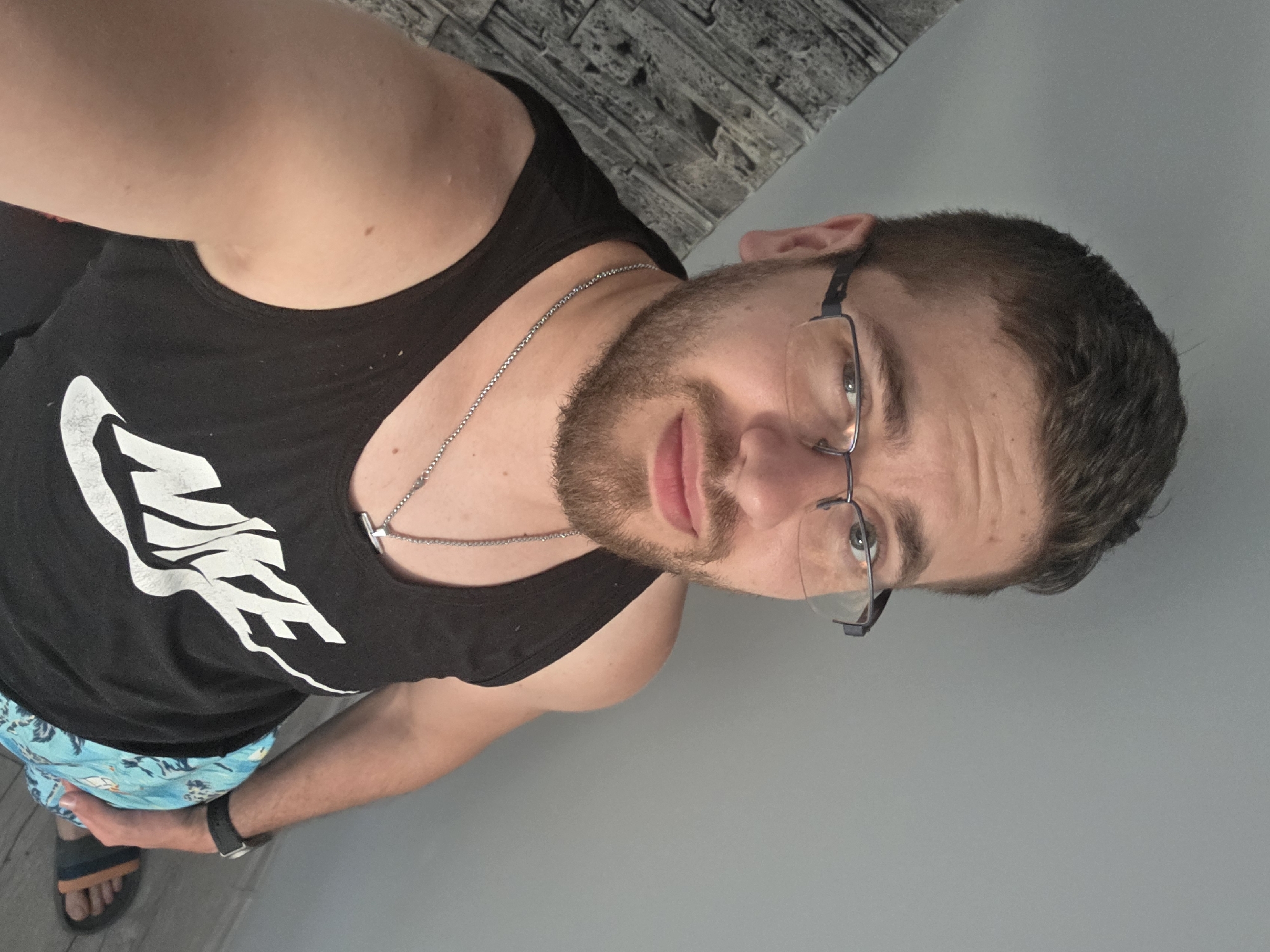 An indoor selfie, taken from a slightly low angle, depicts a male individual in Yagodovo, Bulgaria. The subject, appearing to be between 25 and 35 years old, has short, dark brown hair with faded sides, a trimmed brown beard, and a fair complexion. He wears dark-rimmed rectangular glasses and looks directly into the camera. His attire consists of a black tank top featuring the white "NIKE" logo on the chest, paired with light blue patterned shorts. Accessories include a silver chain necklace with a rectangular pendant, a black watch on his left wrist, and blue and orange flip-flops on his feet. The background on the right and top is a plain light grey wall. On the left, a diagonally ascending textured grey wall, resembling stone or brick, is visible. The floor in the lower right corner is light-colored wood or laminate. The scene is evenly illuminated.