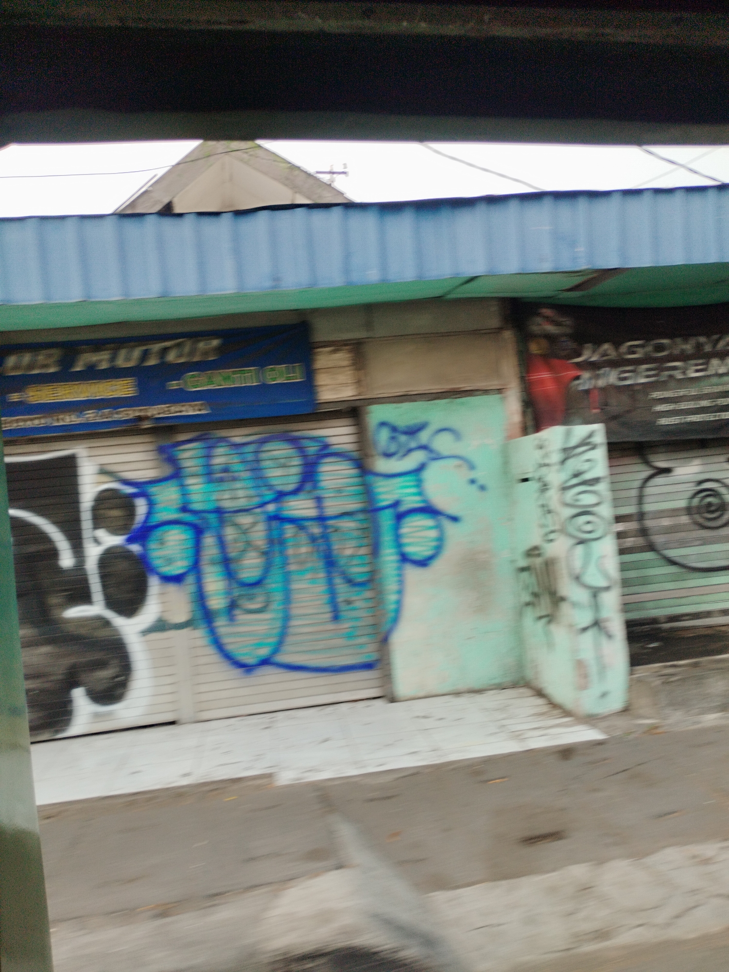 This image captures a street scene in Surabaya, Indonesia, likely a commercial area. In the foreground, a blurry concrete surface suggests a road or sidewalk. Behind this, a series of closed shop fronts with rolling metal shutters dominate the midground. Some of these shutters are covered in graffiti, notably a large, stylized blue and white tag on the left and a lighter blue graffiti piece on the shutter in the center. A green pillar is visible on the right side, partially obscuring another shutter with graffiti. Above the shops, a blue corrugated awning runs horizontally, with glimpses of a building's roof structure peeking out from behind it. A dark, blurry band at the top of the image likely represents an overpass or structure casting a shadow. The overall impression is one of urban decay and vibrant street art in a commercial district.