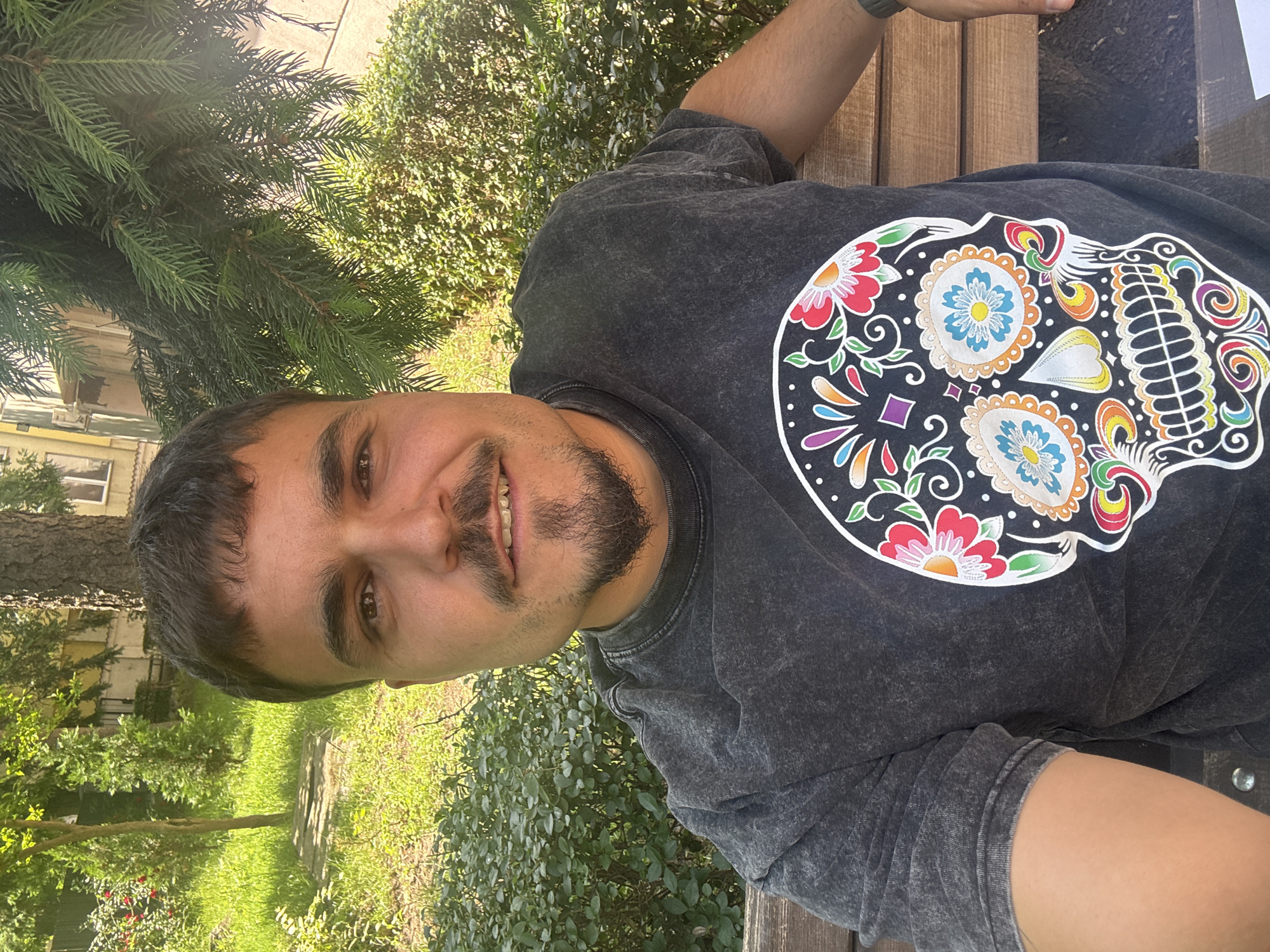 A man is lying on a wooden bench in a park. He is wearing a dark-colored t-shirt with a colorful sugar skull design. The park has green grass and trees. A building is visible in the background. The photo was taken in Plovdiv, Bulgaria.