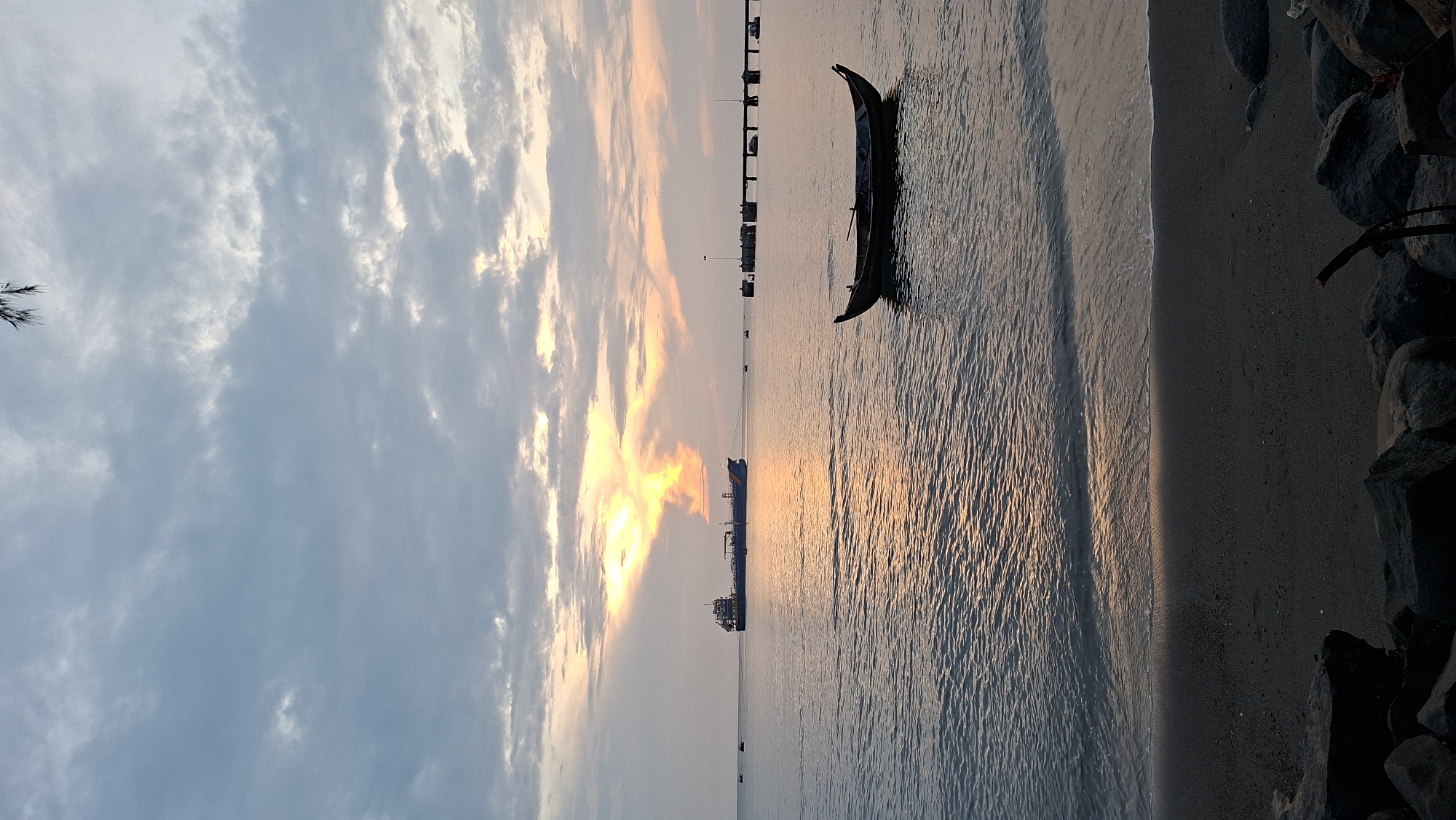This is a vertical, eye-level shot of a tranquil ocean scene at dusk, likely taken from a beach. The foreground shows dark rocks and a sandy shore where gentle waves are washing ashore. The midground is dominated by the calm sea, reflecting the warm hues of the setting sun, which is breaking through a cloudy sky. A small, dark wooden boat is anchored in the water on the right side of the frame. Further out in the water, a large blue and white cargo ship is visible on the left, tethered to a distant pier or dock structure with a row of pylons. The sky is filled with a dramatic display of clouds, illuminated from behind by the setting sun, creating streaks of orange and yellow light. The overall atmosphere is serene and peaceful, capturing the beauty of twilight over the ocean. This scene is characteristic of the coastal areas in Lhokseumawe, Indonesia, known for their harbors and maritime activity.