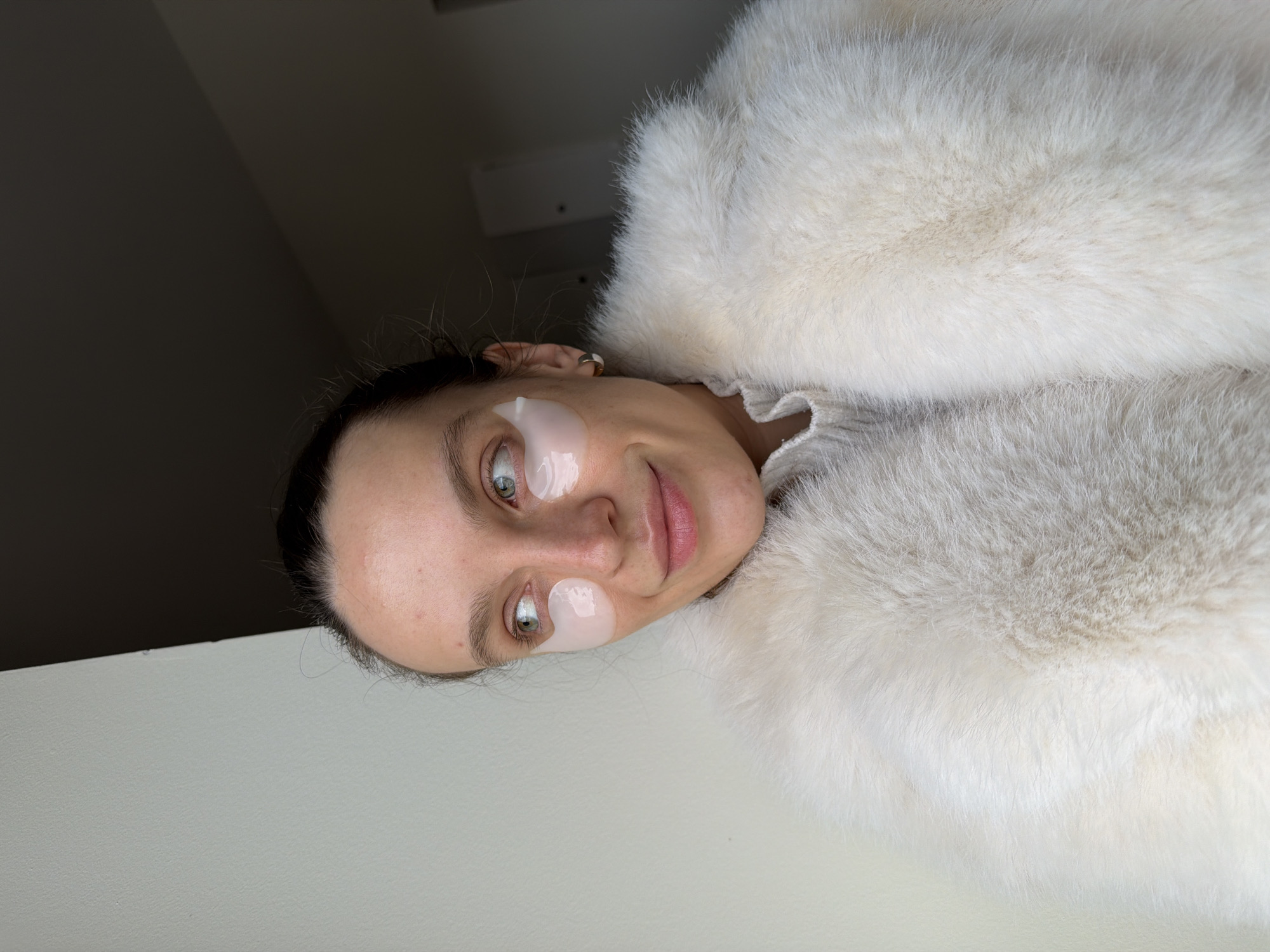 An adult female is depicted indoors, situated in Budva, Montenegro. She is positioned in a medium close-up, facing slightly left with her head turned to look towards the upper left quadrant of the frame. Her hair is pulled back, revealing a fair complexion, light-colored eyes, and a slight, closed-mouth smile. Two translucent, light-colored gel eye masks are applied beneath her eyes. A small, gold-toned hoop earring is visible in her right ear. She is dressed in a light-colored, ribbed knit top, partially visible at the neck, underneath a prominent cream-white fur coat. The background consists of a plain, light-colored wall on the left and a darker, shadowed wall or area on the right, indicating an interior setting with natural or ambient lighting from the left.