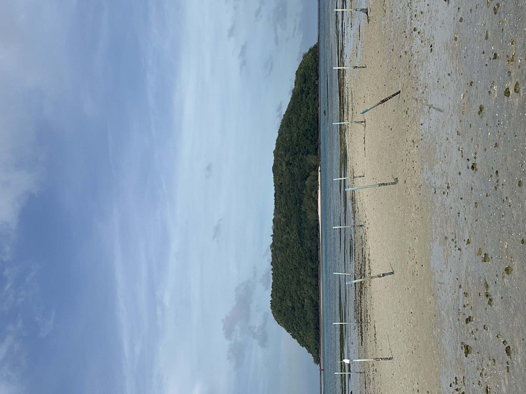 The image depicts a tropical island landscape under a partially cloudy sky. In the foreground, a sandy beach is wet with shallow water and scattered with rocks and shells. Several wooden posts, some capped with blue, are planted in the sand, creating a linear pattern. The middle ground features calm, blue water with submerged structures visible beneath the surface. In the distance, a large, verdant island rises from the sea, covered in dense green foliage. The overall scene suggests a coastal area in a tropical region, possibly a shallow bay or estuary at low tide. The location is specified as Koh Phangan Sub-district, Thailand.
