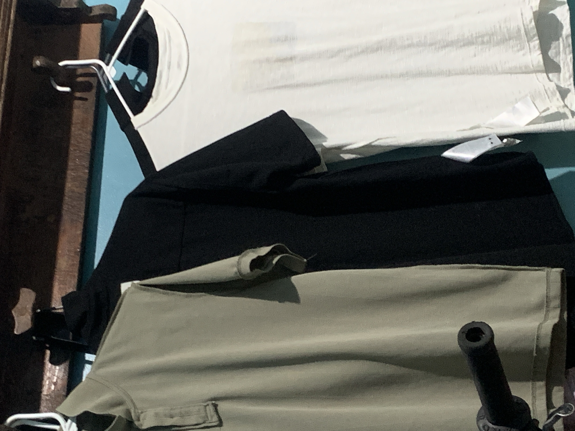 Three t-shirts are hanging on hangers on a wooden shelf against a blue wall. From left to right, the t-shirts are olive green, black, and white. The olive green t-shirt is a polo shirt with a collar and buttons. The black t-shirt is a crew neck. The white t-shirt is also a crew neck and has a tag attached. A black motorcycle handlebar is visible in the lower left corner of the image. The scene is likely indoors, possibly in a closet or dressing area. No specific location in Santa Ana, Venezuela, is identifiable from the image.