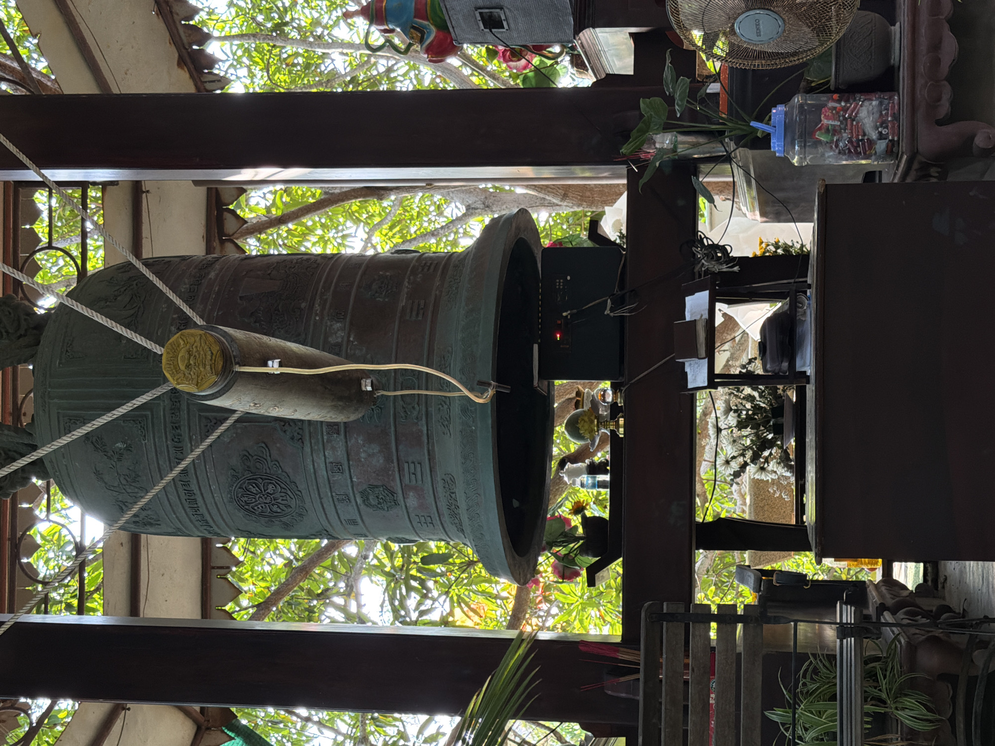 The image shows a large, dark green bronze bell hanging from a wooden structure in what appears to be a temple or shrine. The bell is intricately decorated with patterns and inscriptions. A thick rope is attached to the top of the bell, used for striking it. Below the bell, there is a dark electronic device with a red light visible. Various objects are placed on wooden shelves and stands around the bell, including plants in pots, sticks of incense, and decorative items. A pedestal with a small statue of a seated figure with a colorful headdress is visible to the right, next to a speaker. In the foreground, there is a wooden bench with plants and a dark wooden cabinet. The background is filled with lush green trees and foliage, suggesting an outdoor or semi-outdoor setting. The overall atmosphere is serene and spiritual, indicative of a place of worship. Based on the architecture, ornamentation, and the presence of Buddhist iconography, this scene is likely located at a temple in Nha Trang, Vietnam.