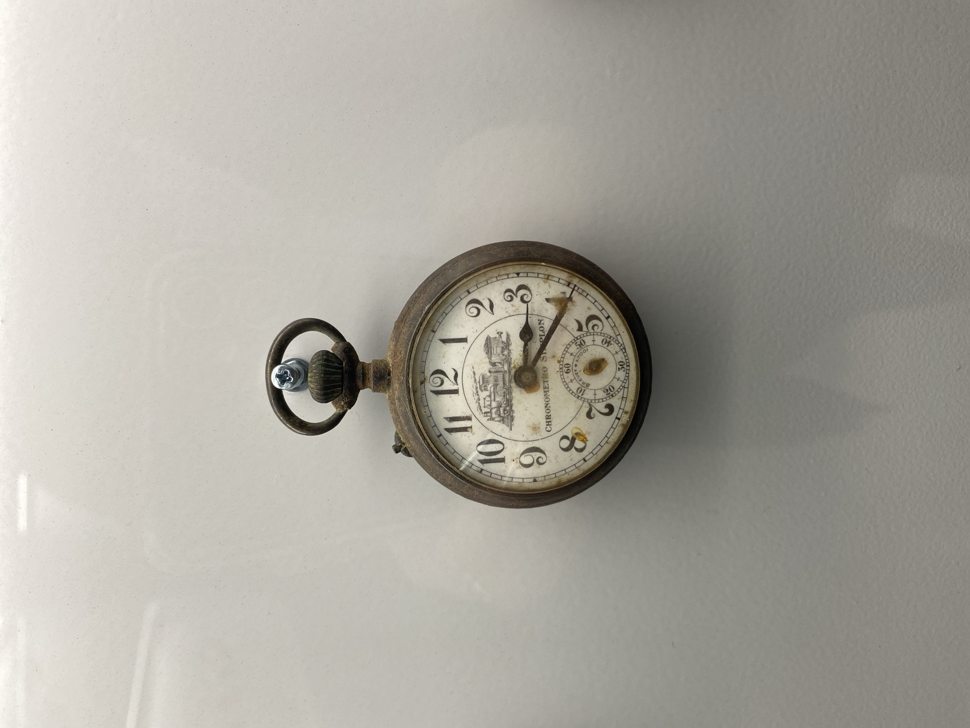 The image shows an antique, possibly non-functional, pocket watch resting on a reflective white surface. The watch has a tarnished metal casing and a white face with black numerals and markings. A small illustration of a train is visible on the lower part of the face. The crown of the watch is fitted with a screw. The time is approximately 1:18. The watch brand is "Chronometre s/bron". The location of the watch in relation to the city of Gijón/Xixón, Spain is undetermined. There are no people depicted in the image.