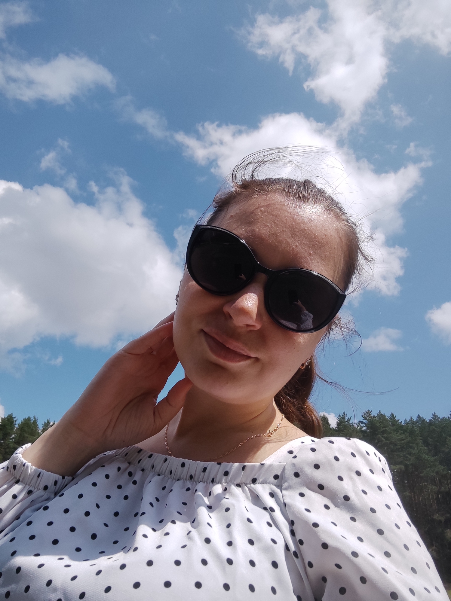 A woman is captured in a close-up, low-angle selfie, holding her hand to her neck. She is wearing large, black sunglasses and a white off-the-shoulder top with black polka dots. Her hair is pulled back, with wisps of it blowing in the wind. The background consists of a bright blue sky with scattered white clouds, and a dense line of evergreen trees can be seen in the distance. The image appears to have been taken outdoors on a sunny day.
