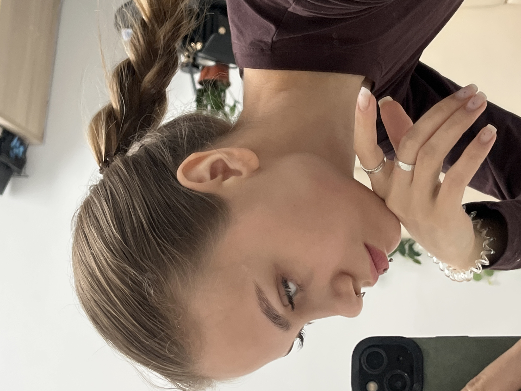 A woman is pictured indoors. She has blonde hair styled in a braid and is wearing a maroon long-sleeved shirt. The woman has several piercings on her lip and nose. She is wearing two rings on her hand, and her nails are manicured with white tips. Her hand is resting on her face. She is holding a cell phone. The specific location is unknown.