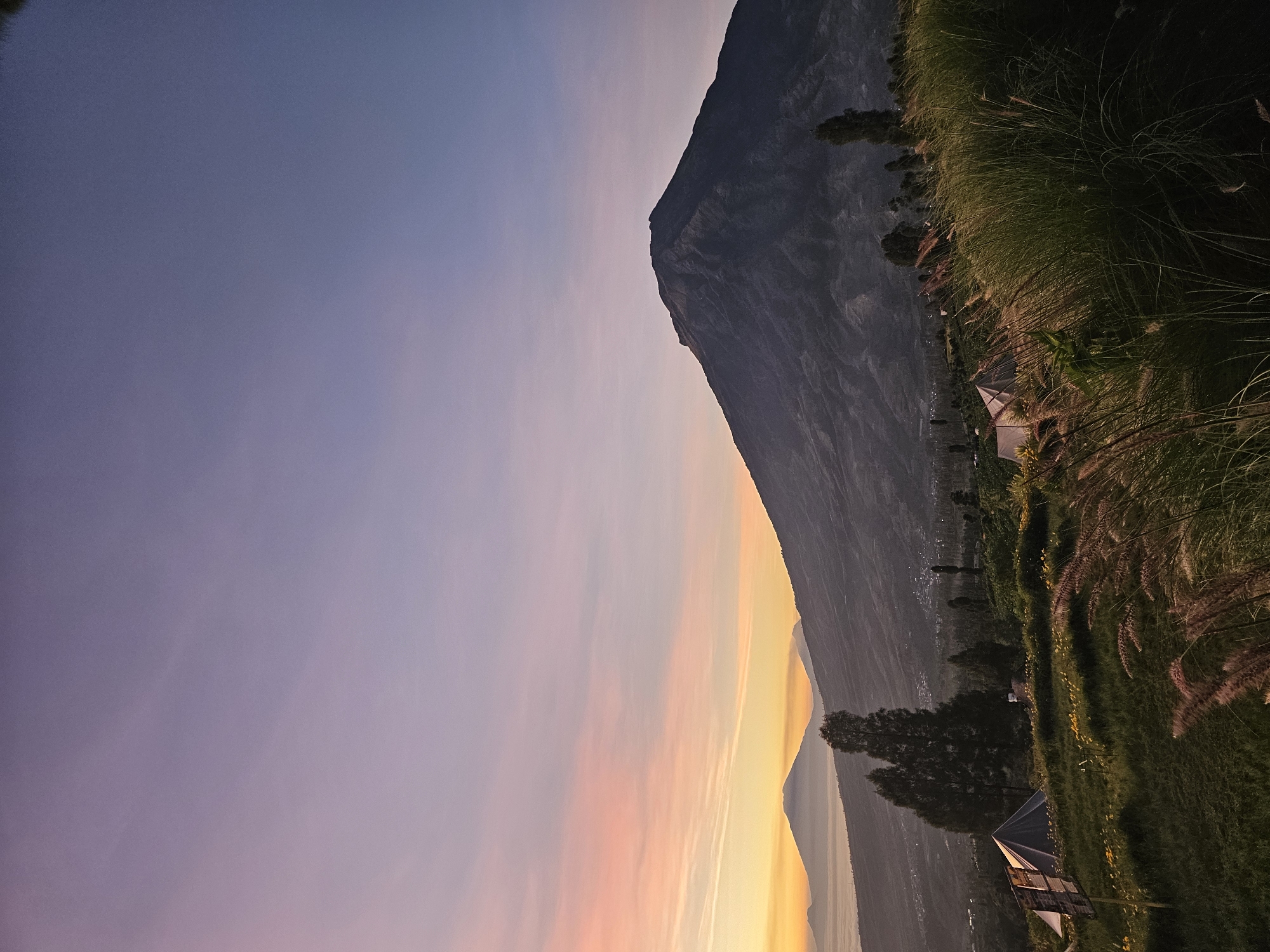 A wide-angle, eye-level, outdoor, vertical, photograph depicts a serene landscape at dawn, likely in Tlahab, Indonesia, characterized by a dramatic sky and mountainous terrain. The sky transitions from a soft pink and orange hue near the horizon to a deep violet and blue at the zenith. In the foreground, tall, wispy grasses are in motion, suggesting a gentle breeze. Scattered throughout the middle ground are several camping tents, some dark and triangular, others lighter and dome-shaped. Some yellow flowers are visible amongst the green grass.
Dominating the right side of the frame is a large, dark, volcanic mountain, its slopes rendered in shadow with subtle texture. To the left, a distant mountain range is partially obscured by a layer of soft mist or clouds near its base, with a hint of a town or village nestled in the valley below. The overall atmosphere is tranquil and natural, indicative of an early morning in a rural, mountainous region. There are no people visible in the frame.