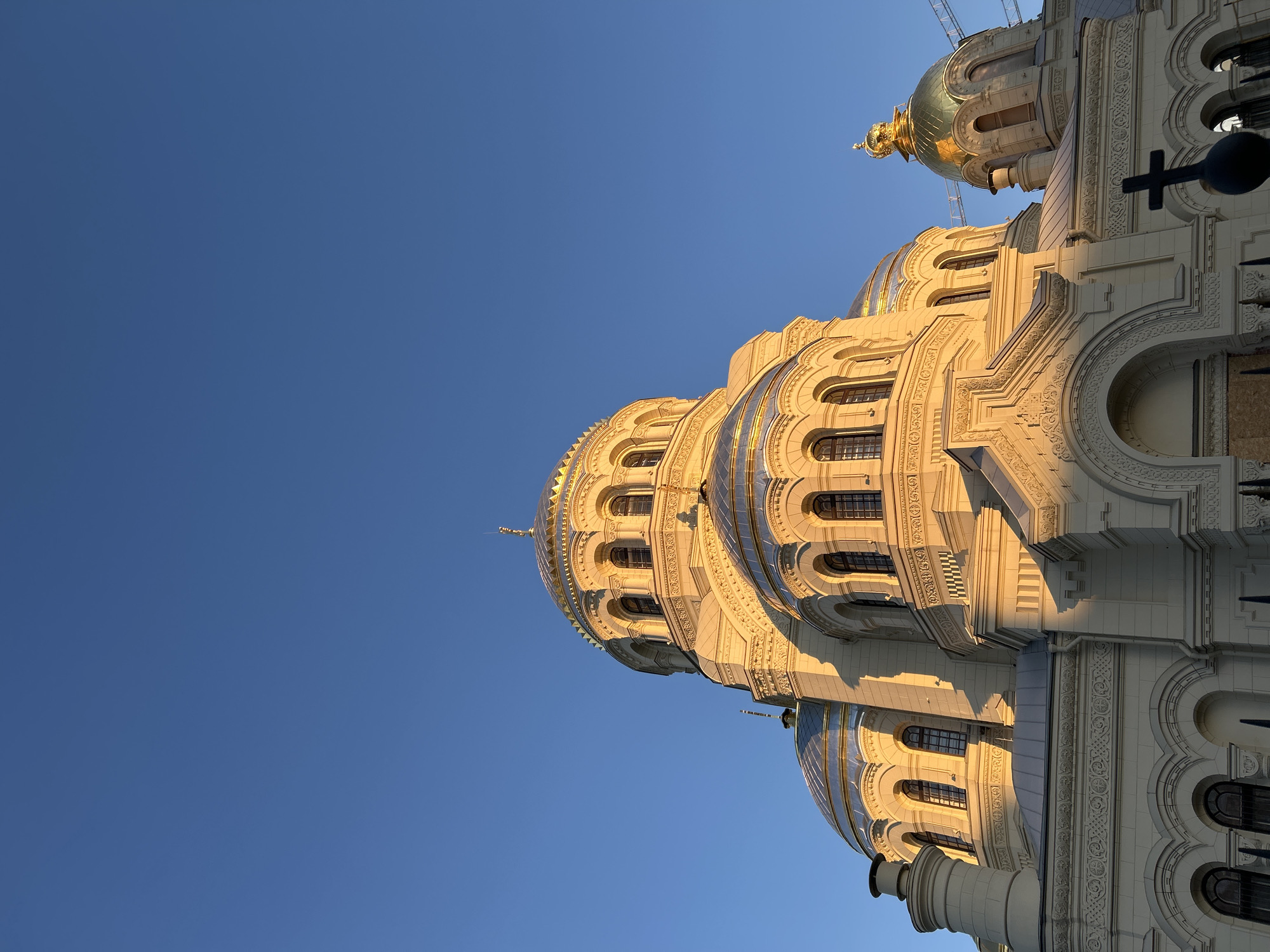 The image shows the Naval Cathedral of Saint Nicholas in Kronstadt, Saint Petersburg, Russia, from a low angle. The cathedral's architectural details are highlighted by the sunlight, with golden domes visible against a clear blue sky. The façade is ornate, featuring windows and decorative arches. A cross is partially visible on the right side of the frame. The presence of scaffolding suggests that the building is undergoing construction or renovation.