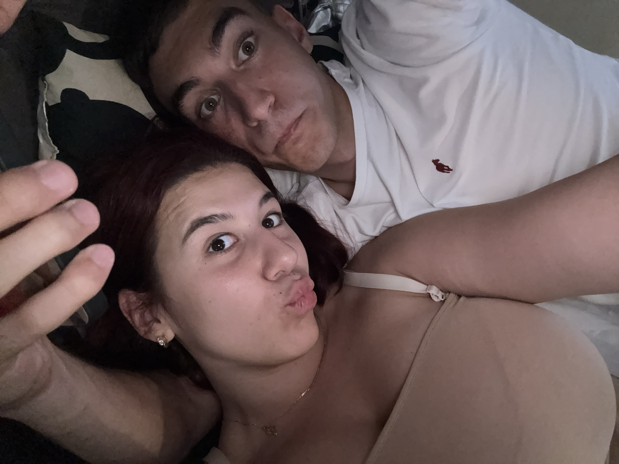 A couple is lying down together in an indoor environment. The woman, with dark hair, is in the foreground, looking at the camera with pursed lips. She is wearing a beige top and a necklace. Behind her, a man is looking at the camera with a neutral expression, wearing a white polo shirt with a red logo. The environment appears to be a casual, relaxed setting. The location is Unknown, Unknown.