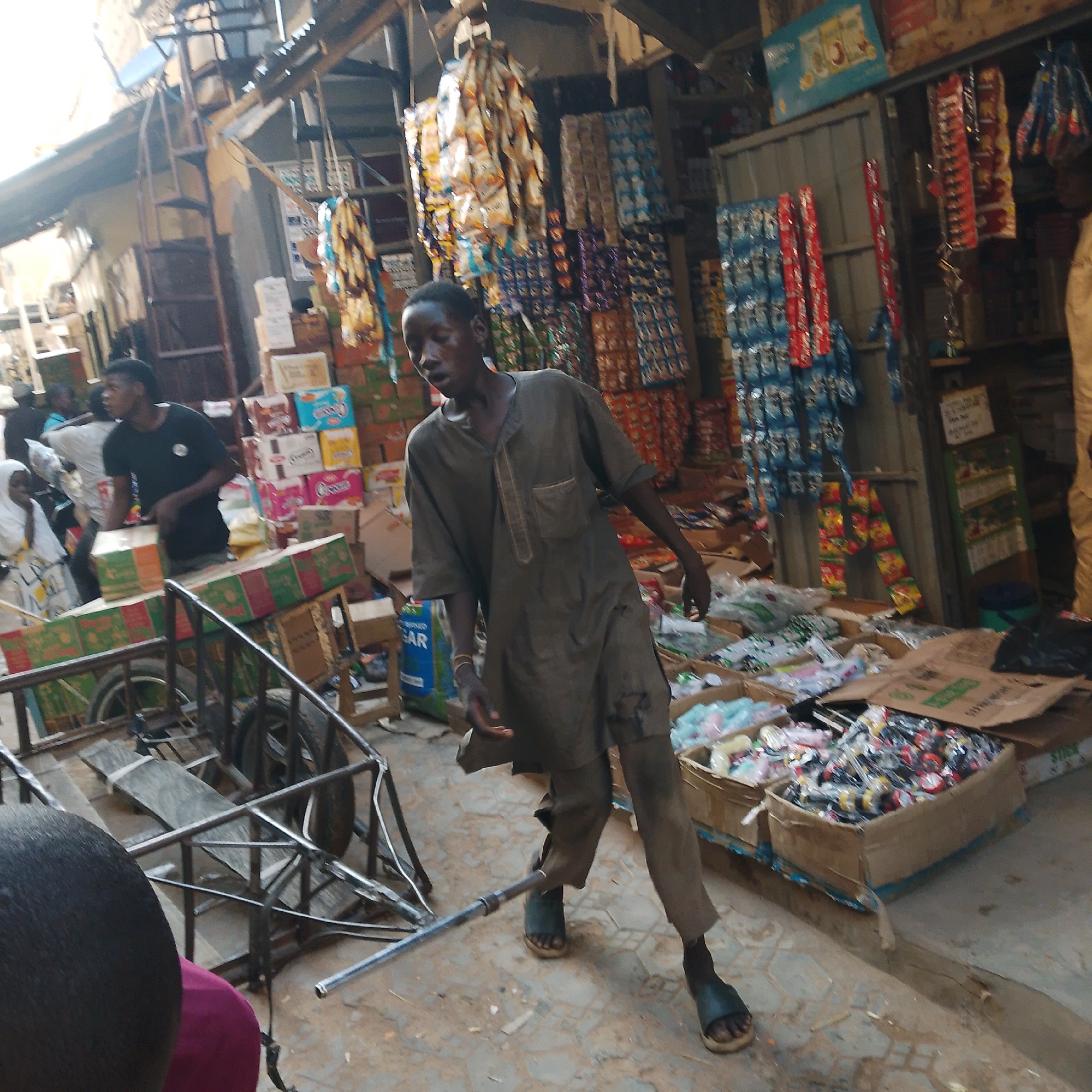 The image depicts a bustling marketplace scene in Arawa, Nigeria. A young man wearing a dark brown, traditional tunic and pants is the central figure, walking and pushing a handcart laden with green and red boxed goods. His gaze is directed downwards, and his left hand is on the handle of the cart. Beside him, another person in a black t-shirt is visible, looking towards the left. In the background, a woman in a white headscarf and patterned dress is partially obscured. The marketplace is filled with stalls displaying various packaged goods, including brightly colored snacks and drinks. Cardboard boxes are stacked high, suggesting a busy commercial environment. The ground is paved with earth-toned tiles. The overall impression is one of vibrant market activity in a Nigerian city.