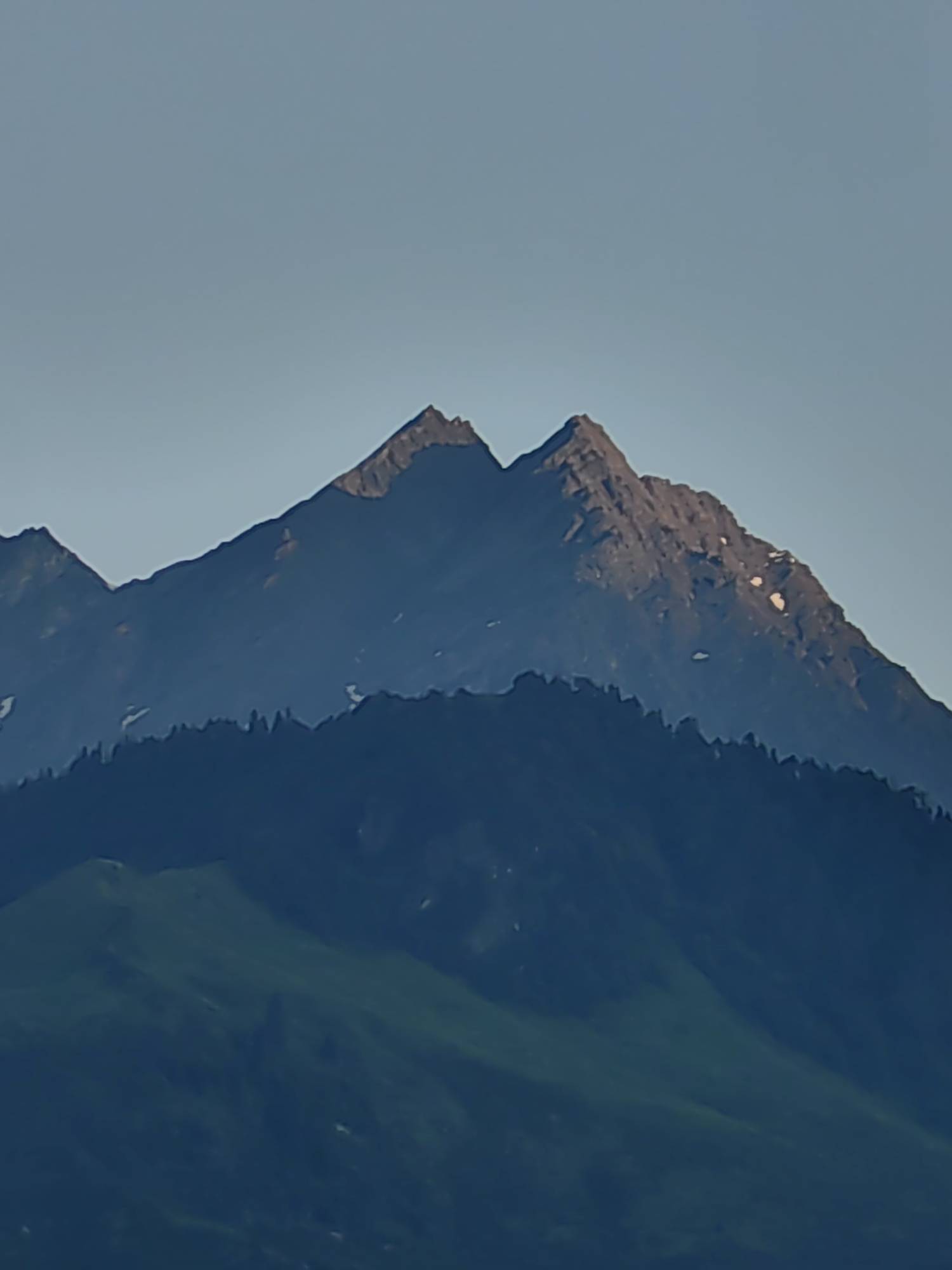 The image displays a mountain range under a clear, pale blue sky. The mountains are layered, with the foreground mountains appearing darker and more rounded, covered in green vegetation and trees. The middle ground features a darker, more densely forested mountain slope. The background reveals sharp, rocky peaks, with snow patches visible on the uppermost mountain. The lighting suggests it is either early morning or late afternoon, casting a soft glow on the peaks. The scene is devoid of any human presence or man-made structures. The location in Vashisht, India, implies this is a view of the Himalayan foothills.
