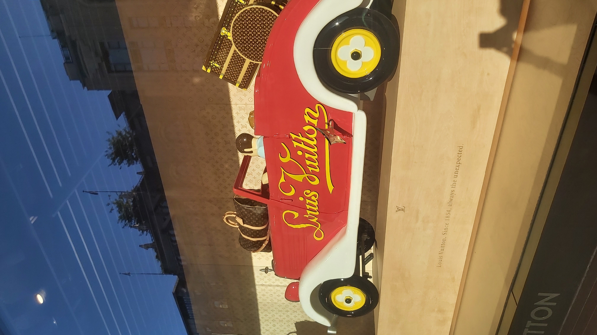 This image shows a red toy car with "Louis Vuitton" written in yellow script on its side, displayed in a store window. The car has two Louis Vuitton suitcases on top and in the back. A Louis Vuitton logo is visible on the background wallpaper and on a wooden platform below the car. Text on the platform reads, "Louis Vuitton. Since 1854, always the unexpected." The reflection on the window shows a building, trees, and flags, suggesting it is an outdoor display. The specific location in Paris, France, cannot be determined from the image.