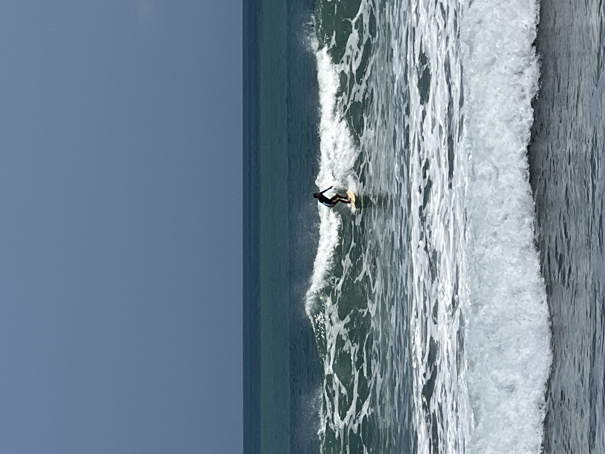 A lone surfer, wearing a dark wetsuit, rides a small wave in the ocean off the coast of Kuta, Indonesia. The surfer is positioned on a light-colored surfboard, leaning forward as they navigate the wave. The water is a deep teal color, with white foam cresting the wave and scattered across the surface. The sky above is a muted, overcast blue, with a darker horizon line where the sky meets the sea. The image captures a moment of active recreation in a coastal environment.