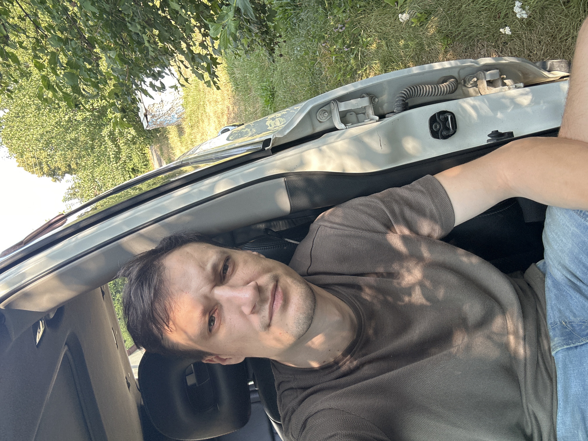 A man is sitting in the driver's seat of a car, with the driver's side door open. He is wearing a brown t-shirt and blue jeans. The car is parked in a grassy area with trees in the background. The sun is shining, casting dappled shadows on the man and the car. The scene appears to be in a rural area, possibly on the outskirts of Shatava, Ukraine. There is a small, blue and white structure visible in the distance, partially obscured by foliage. The image is a selfie, taken by the man.