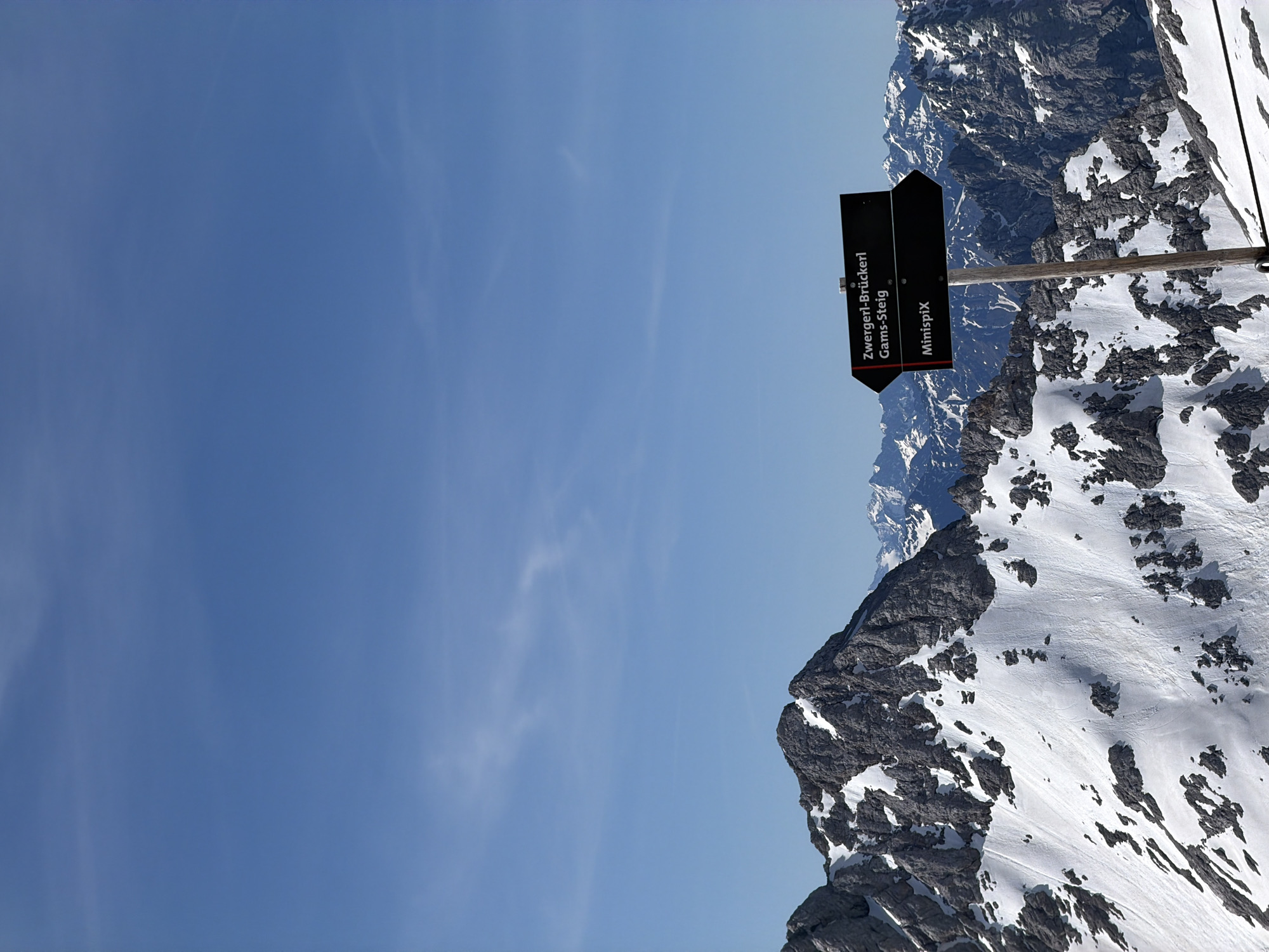 This is a landscape photograph taken on a clear day in the Bavarian Alps, likely near Garmisch-Partenkirchen, Germany, given the German signage. The image features a signpost with two black directional arrows. The top arrow points left and reads "Zwergerl-Brückerl Gams-Steig". The bottom arrow also points left and reads "MinispiX". The signpost is made of wood and has a weathered appearance. Behind the signpost, a vast expanse of snow-covered mountains dominates the scene. The mountains are rugged and rocky, with patches of snow clinging to their slopes and peaks. The sky above is a vibrant blue, with a few wispy clouds. The overall impression is one of a high-altitude alpine environment, likely a popular hiking or skiing destination. There are no people or animals visible in the image.