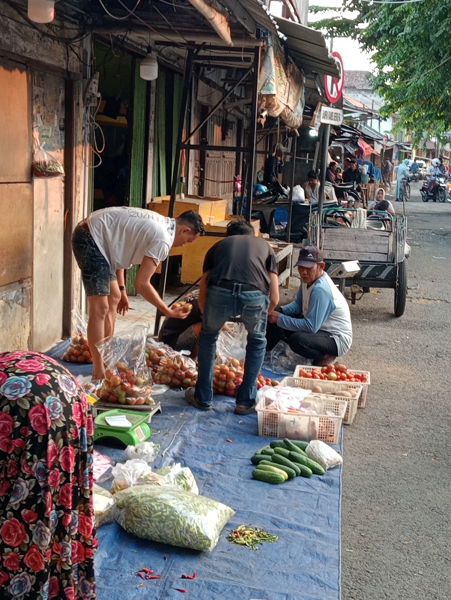 The image depicts a street market scene in Demak, Indonesia, likely in the morning or late afternoon given the lighting. In the foreground, a person wearing a floral skirt stands partially visible, facing away from the camera. A blue tarp covers the ground, displaying various fresh produce. Piles of tomatoes are visible in clear plastic bags, some on a green weighing scale. Several baskets filled with tomatoes and what appear to be potatoes are arranged in a line. A large bag of green beans and a bunch of cucumbers are also present. Some scattered red chili peppers are on the tarp.
In the middle ground, two men are actively engaged. One man, wearing a white t-shirt and denim shorts, is bending over and appears to be handling tomatoes. Another man in a dark t-shirt and jeans is also tending to the produce. A third man, wearing a blue long-sleeved shirt and a dark cap, is squatting near the baskets of tomatoes. Behind them, a wooden cart with large wheels is parked on the street. Other people and market stalls are visible in the background, suggesting a bustling market environment. Vehicles, including motorcycles, are present on the street, indicating it's a public thoroughfare. Trees and buildings line the street, contributing to the urban setting.