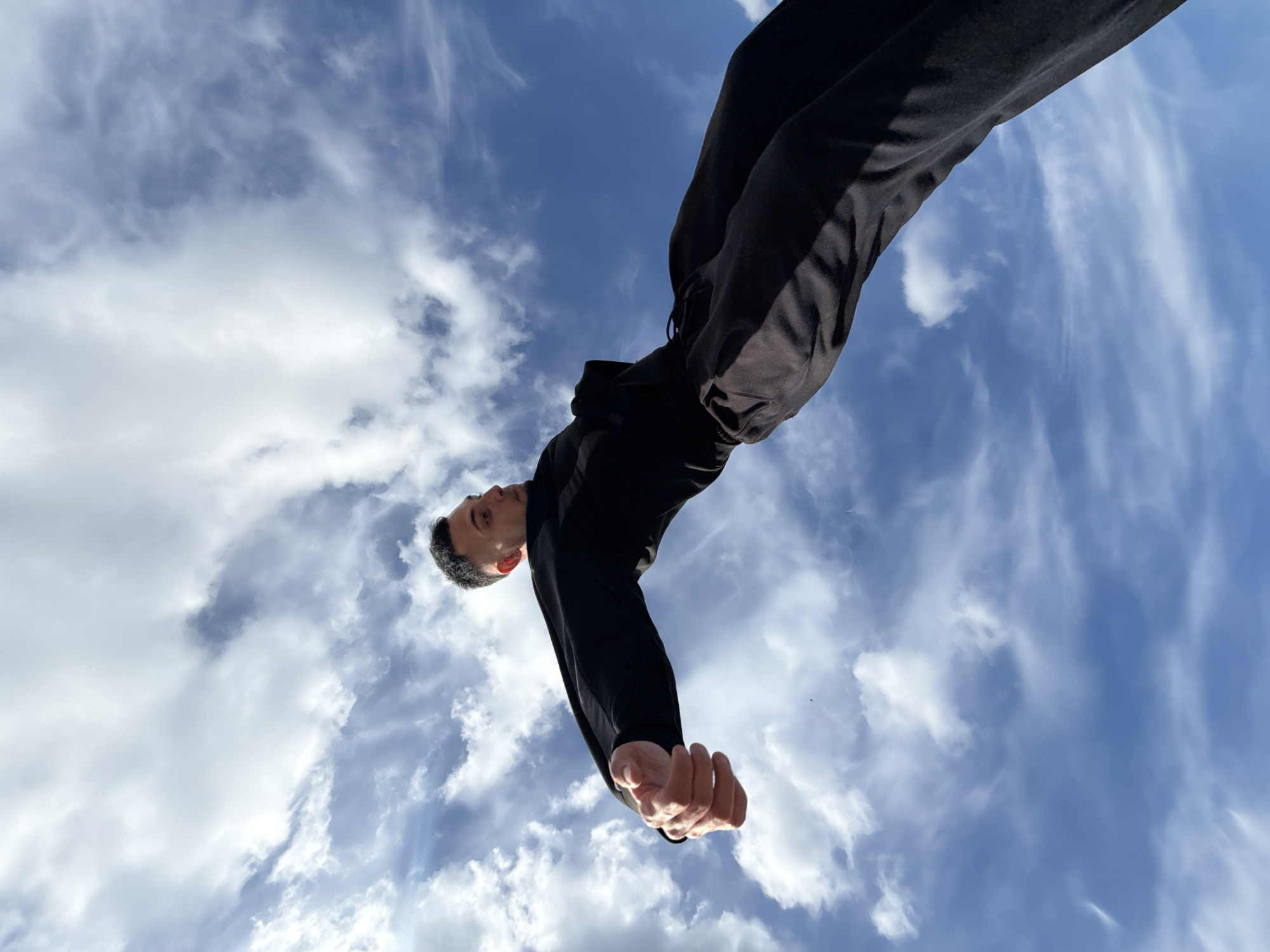 A low-angle shot shows a man in black athletic wear against a sky filled with white clouds and patches of blue. The man is positioned overhead, looking down towards the camera with a neutral expression. His hands are visible and unclenched. The location is an open-air environment, inferred from the sky, but specific details of Shumen, Bulgaria, are absent.