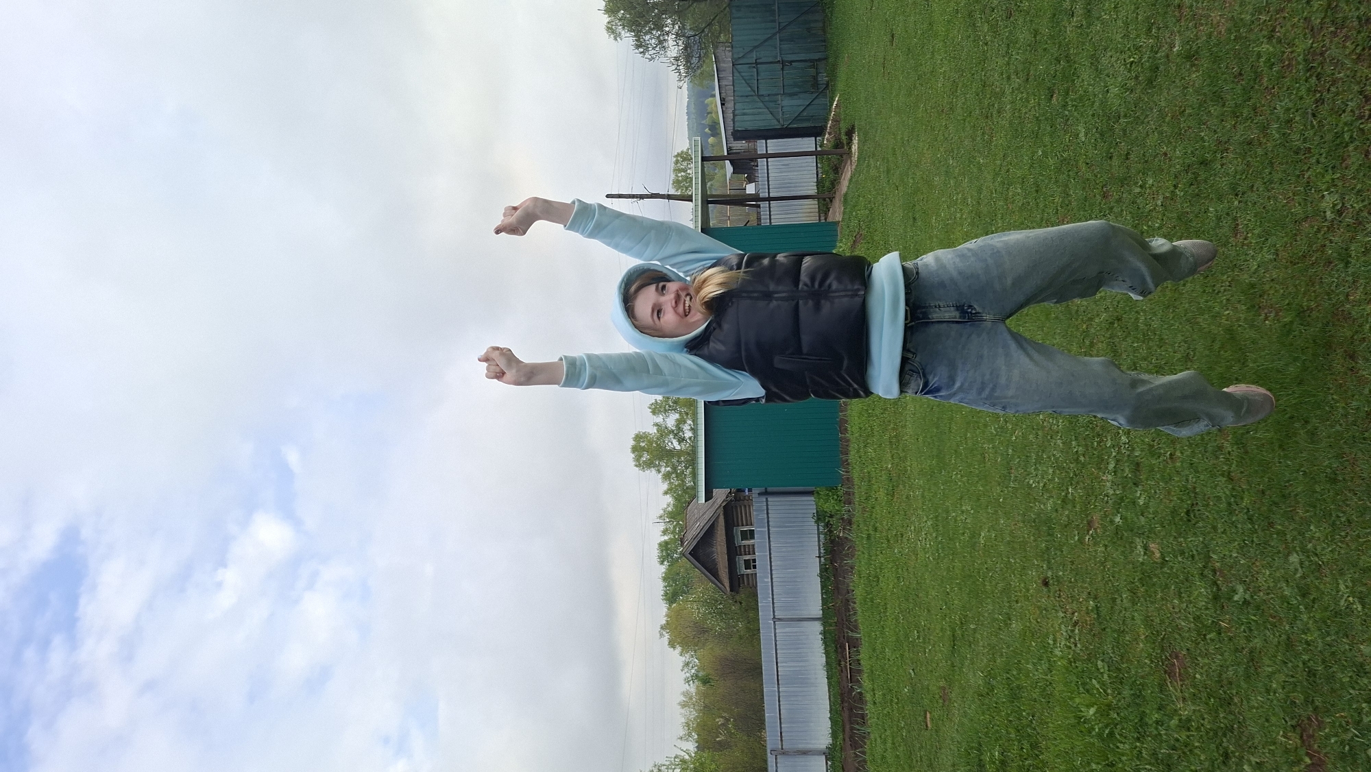 The photograph shows a young woman in a blue hoodie and black vest standing in a grassy area in Большой Букор, Russia. She has her arms raised and appears to be smiling. In the background, there are trees, a fence, and buildings. The sky is cloudy.