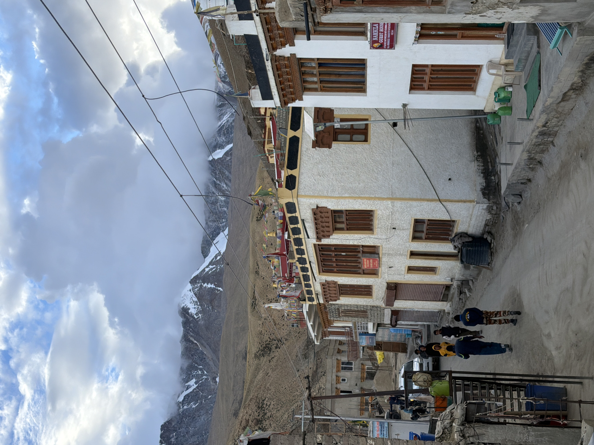 The image depicts a street scene in the village of Karzok, India, situated in a mountainous region. The buildings are predominantly constructed with stucco and wood, featuring Tibetan architectural elements such as prayer flags adorning rooftops and decorative wooden window frames. In the foreground, a paved street runs through the village, with several people walking. Two individuals in the center are walking away from the camera, one wearing a dark blue jacket and camouflage pants, the other in a yellow jacket. A white vehicle is parked on the left side of the street. The background is dominated by rugged, barren mountains, with snow-capped peaks visible in the distance. The sky is partially cloudy, with dramatic cloud formations. The overall impression is of a remote, high-altitude settlement.