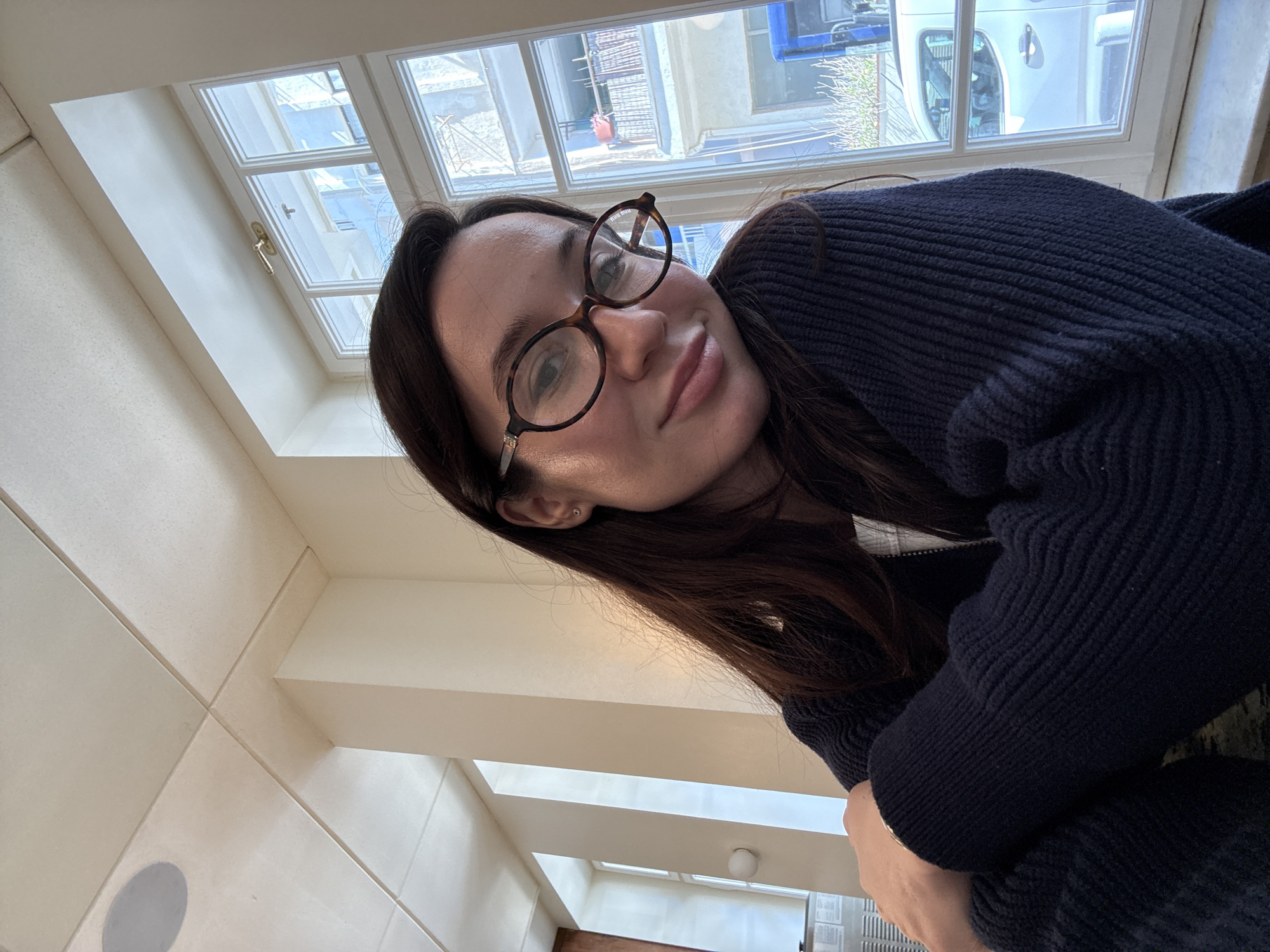 The image is a tilted, close-up selfie of a young woman with long, dark brown hair, looking directly at the camera with a relaxed expression and a subtle smile. She wears large, round, tortoise-shell framed glasses, with the brand "Miu Miu" visible on the right arm. A small stud earring is visible in her left ear, and a thin, light-colored bracelet adorns her right wrist. She is dressed in a dark blue or black ribbed knit sweater or cardigan.
The woman is indoors, positioned in front of a multi-paned window that allows bright natural light to illuminate the scene, indicating it is daytime. Through the window, elements of an urban environment are visible, including parts of other buildings and a large white vehicle or train car. The interior features light-colored walls, possibly tiled or paneled, with distinct architectural angles and a ceiling-mounted speaker partially visible. The setting is consistent with an indoor space overlooking a street or courtyard in an urban area like Tel-Aviv, Israel.