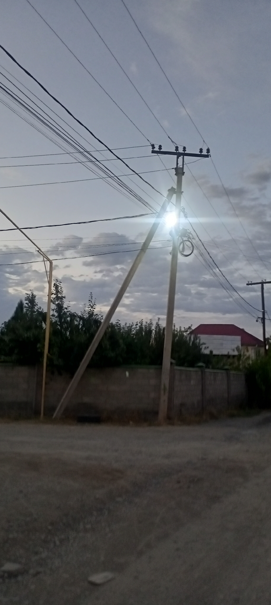 The image depicts a scene with power lines and a leaning utility pole in the city of Romanovka, Kyrgyzstan. The pole, made of concrete and wood, is significantly tilted, with a wooden crossarm supporting multiple electrical wires. A bright light, possibly a street lamp, is illuminated and positioned near the top of the leaning pole, causing a lens flare that obscures details. Supporting cables run from the pole towards the foreground and off-frame. In the background, a low concrete fence runs horizontally across the image, with dense green foliage and trees behind it. Further back, a building with a red roof is visible. The sky is overcast with gray clouds. The ground in the foreground is unpaved and appears to be gravel or dirt. There are no people visible in the image.