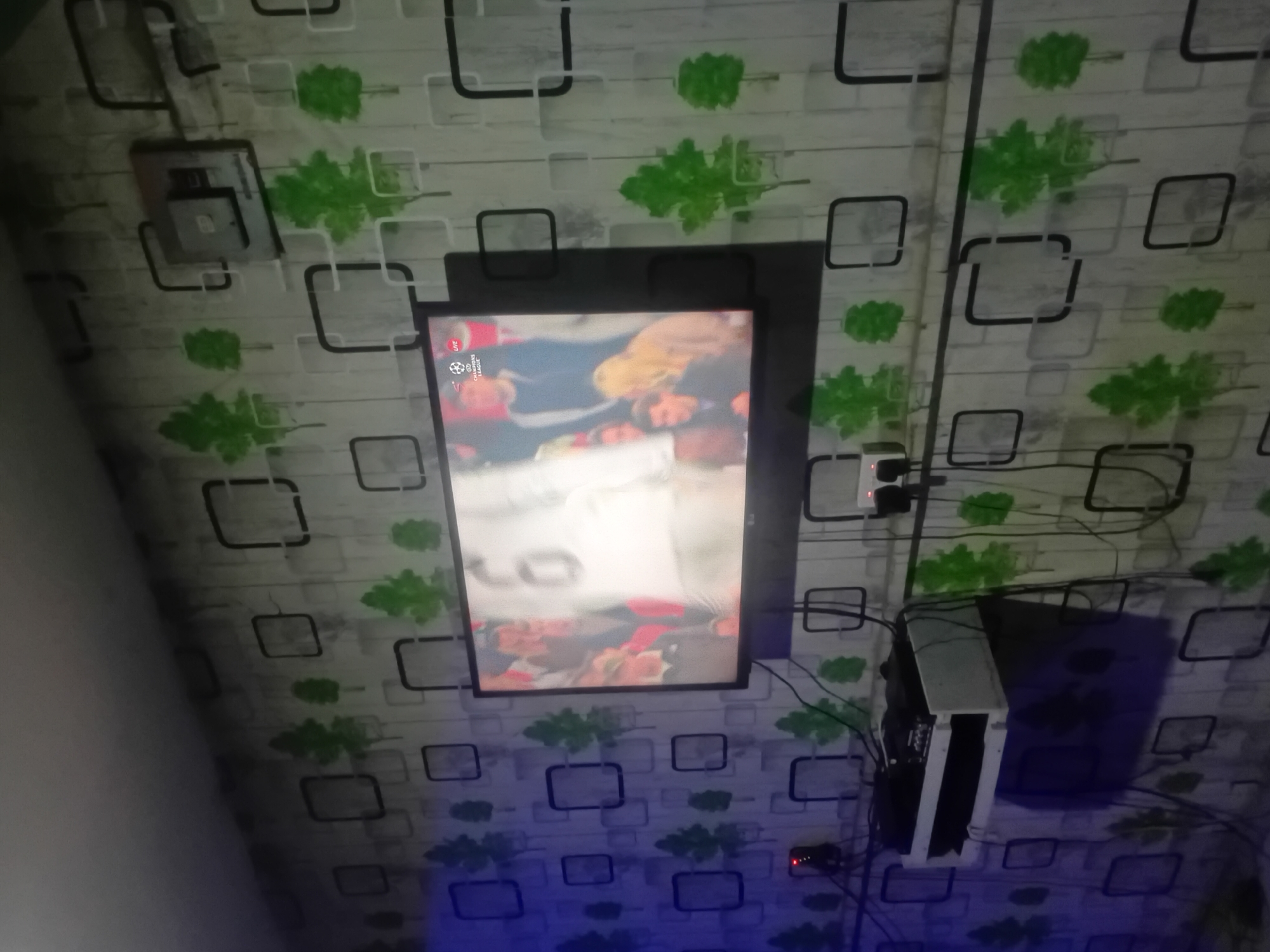 The image shows a monitor mounted on a wall with patterned wallpaper featuring square shapes and green leaf-like designs. Below the monitor, a white computer tower is visible, along with various cables and a power strip. The scene is illuminated with a blue light source at the bottom of the frame. The location is Unknown, Unknown.