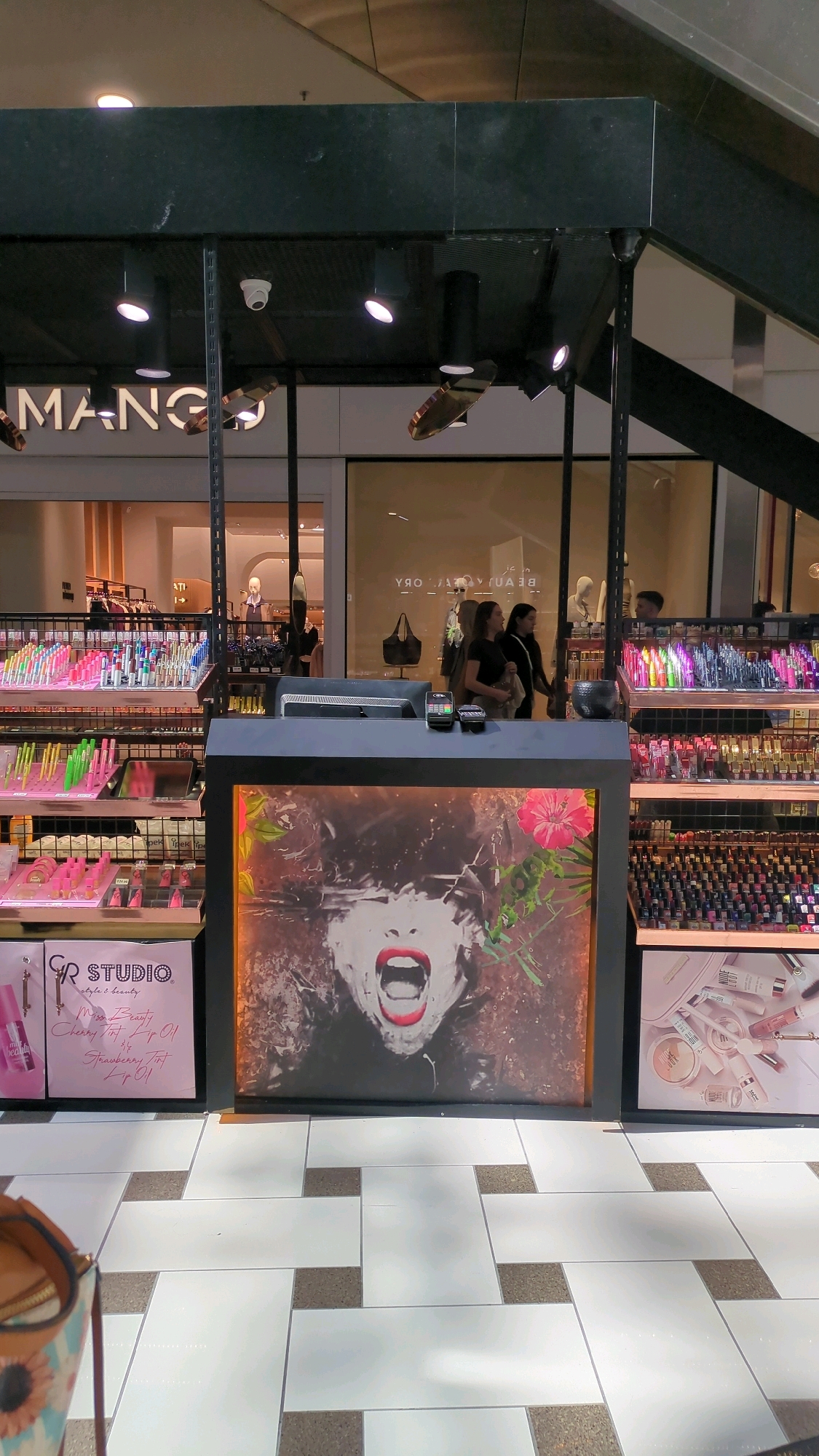 In a brightly lit indoor shopping mall, likely in Chișinău, Moldova, a cosmetics kiosk is prominently displayed. The kiosk features an array of makeup products, including lipsticks, glosses, and nail polishes, neatly arranged on shelves. A striking visual advertisement of a woman's open mouth with bright red lips is displayed on the front of the kiosk, drawing attention. To the right of the kiosk, another advertisement showcases various makeup items. In the background, a store with a prominent "MANGO" logo is visible, suggesting a retail environment within a larger complex. Several people are seen browsing or walking through the mall, with three individuals standing near the "BEAUTY STORY" sign in the background, partially obscured by the kiosk. The floor is tiled in a modern, geometric pattern of white and gray squares. The lighting is a mix of overhead mall lights and spotlights directed at the merchandise.